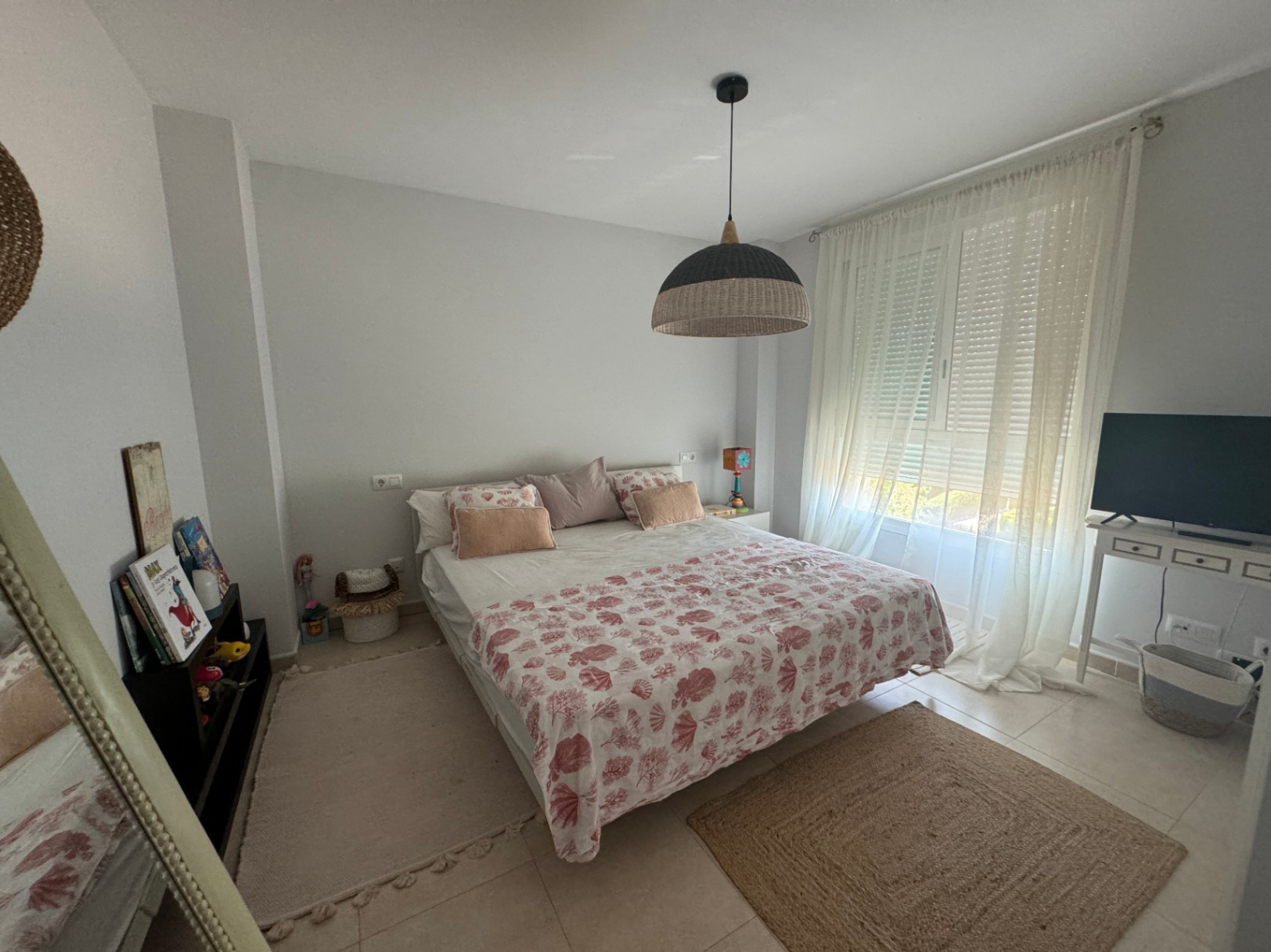 Lichte slaapkamer met tweepersoonsbed, beddengoed met koraalmotief, een geweven hanglamp en een groot raam met witte vitrage.