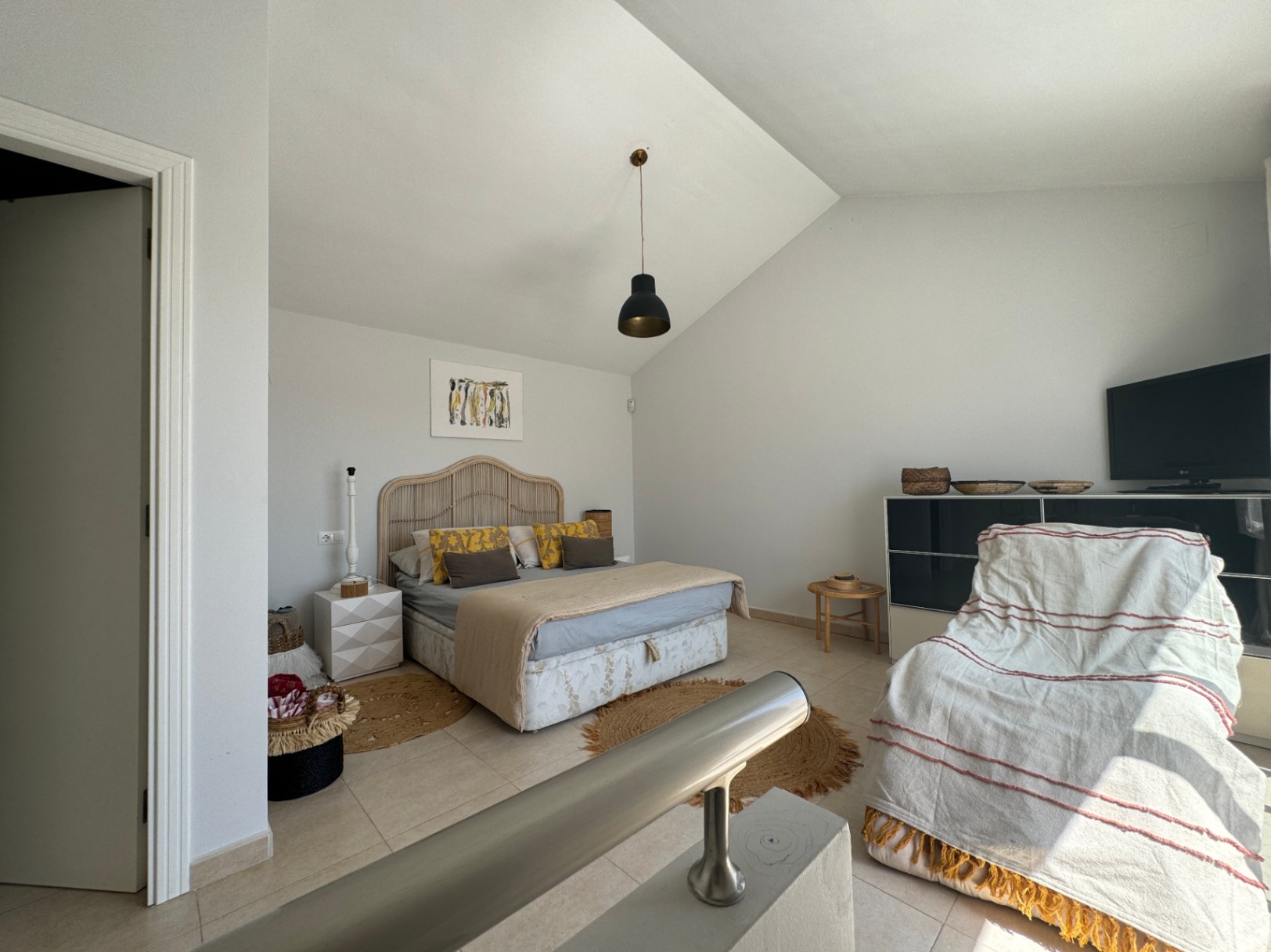 Slaapkamer met schuin plafond, voorzien van een rotan hoofdbord en zwarte hanglamp. De ruimte heeft een tegelvloer, jute kleden en een metalen balustrade op de voorgrond.