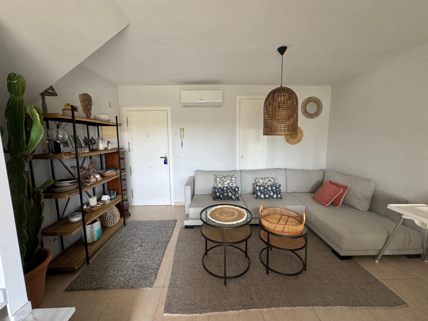 Woonkamer met grijze hoekbank, rotan hanglamp en een industriële houten stellingkast. De lichte tegelvloer en natuurlijke accenten zorgen voor een rustige uitstraling.