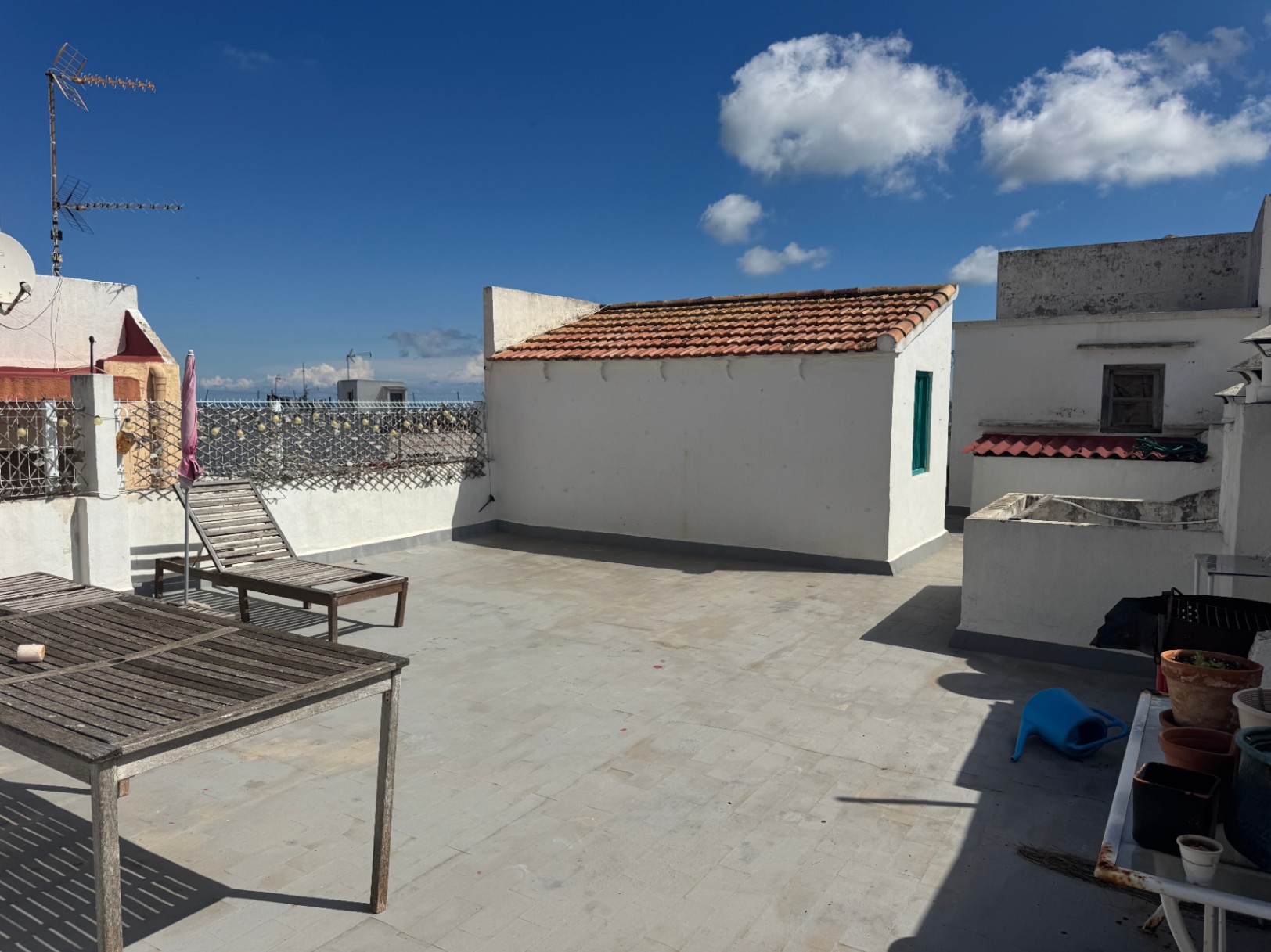 Dakterras met tegelvloer, verweerde houten meubels en een wit bijgebouw met een terracotta pannendak onder een helderblauwe lucht.