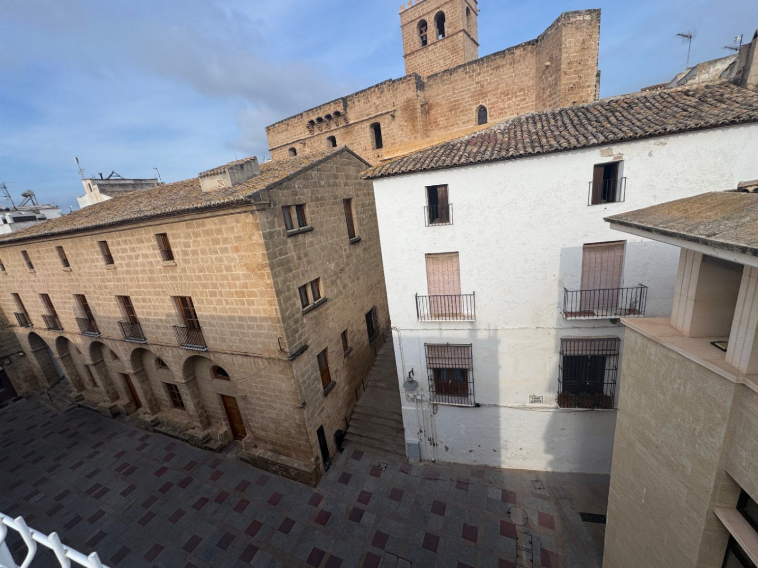 Verhoogd zicht op een historisch plein met stenen gebouwen, witgepleisterde gevels en een karakteristieke kerktoren op de achtergrond.