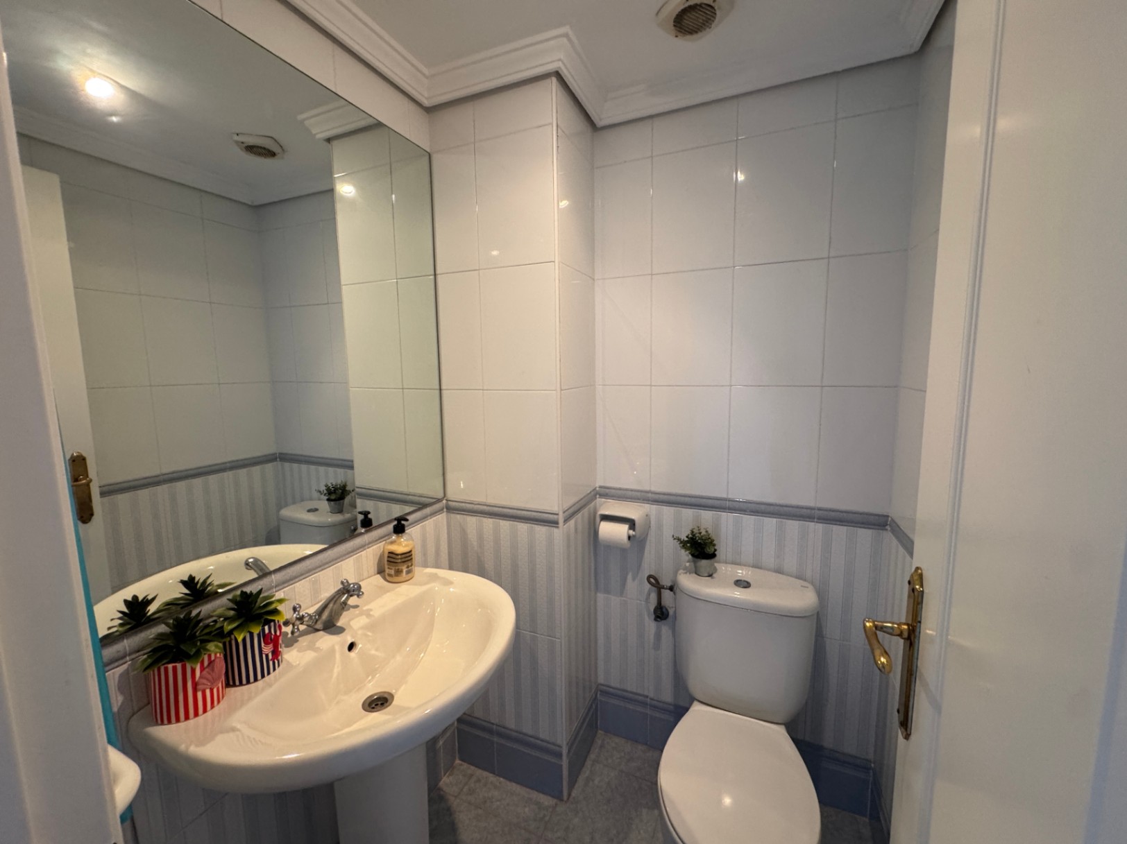 Functionele badkamer met een kolomwastafel, grote spiegel en betegelde wanden met een blauw gestreepte decoratieve rand.
