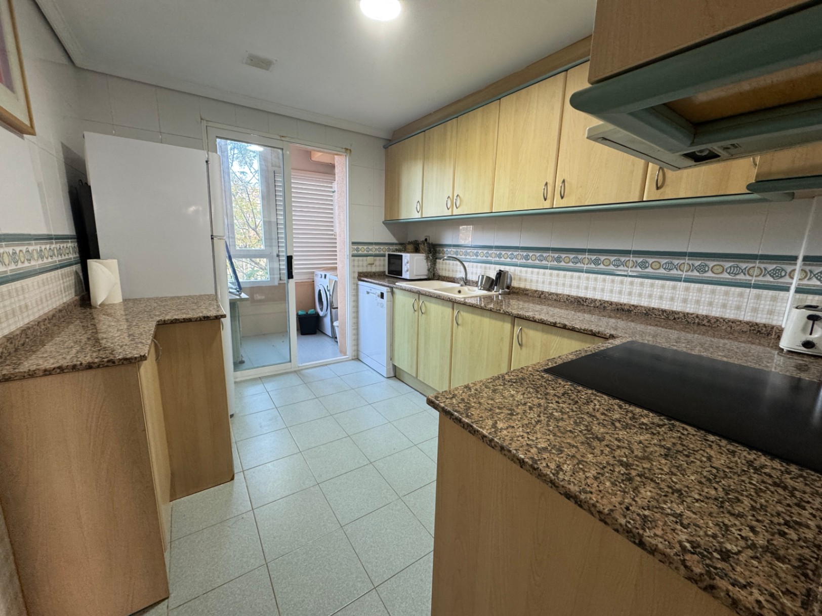 Keuken met lichte houten kasten, granieten werkbladen en toegang tot een bijkeuken op het balkon met een wasmachine via een glazen schuifdeur.