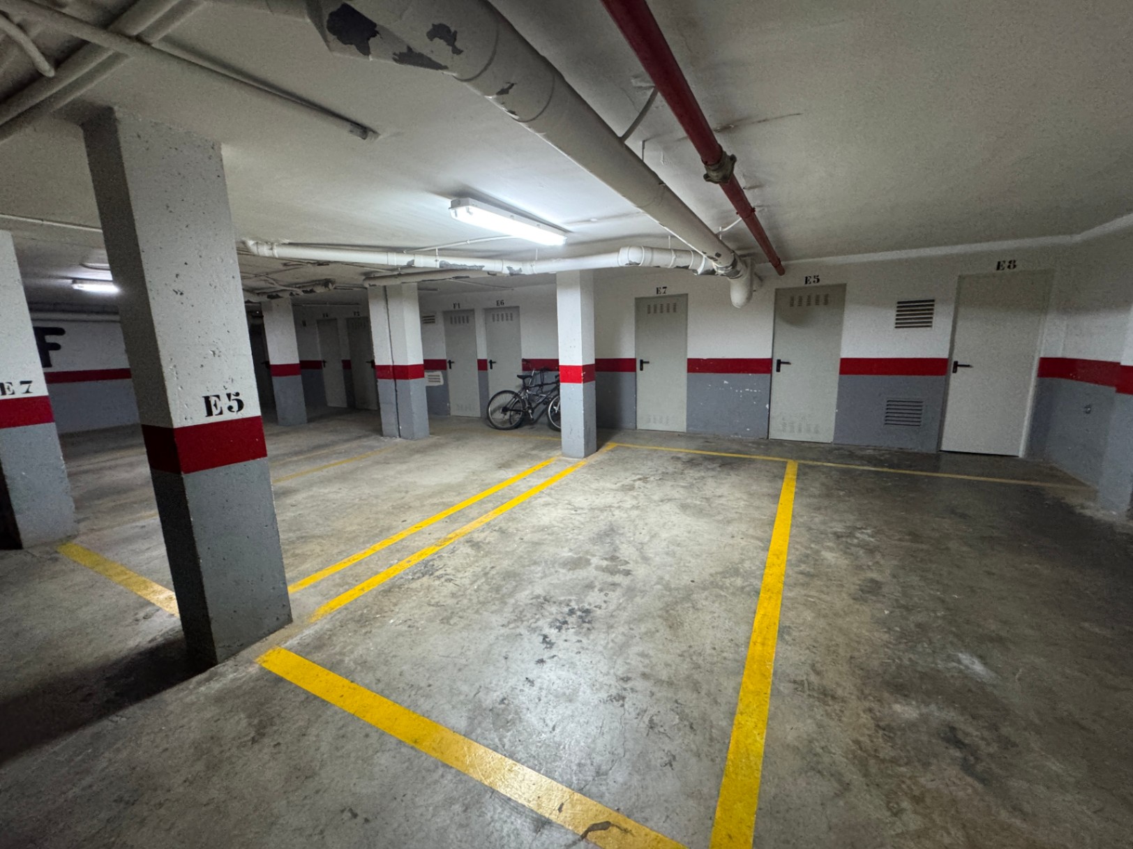 Ondergrondse parkeergarage met betonnen vloer, gele belijning en genummerde bergingen. Voorzien van zichtbare plafondleidingen en functionele verlichting.