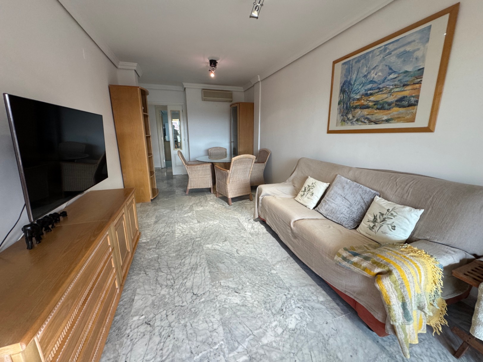 Woonkamer met grijze marmeren vloer, houten tv-meubel en een eethoek met een glazen tafel en vier rieten stoelen op de achtergrond.