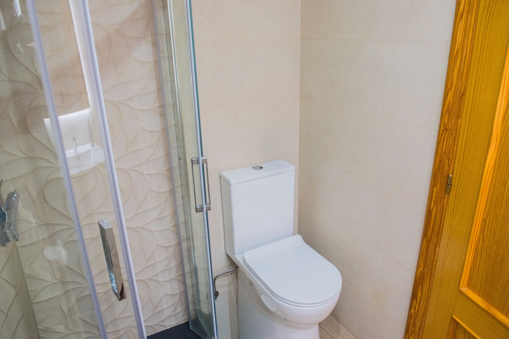 Badkamerhoek met wit toilet en glazen douchecabine, voorzien van decoratieve wandtegels met reliëf en chromen deurgrepen.