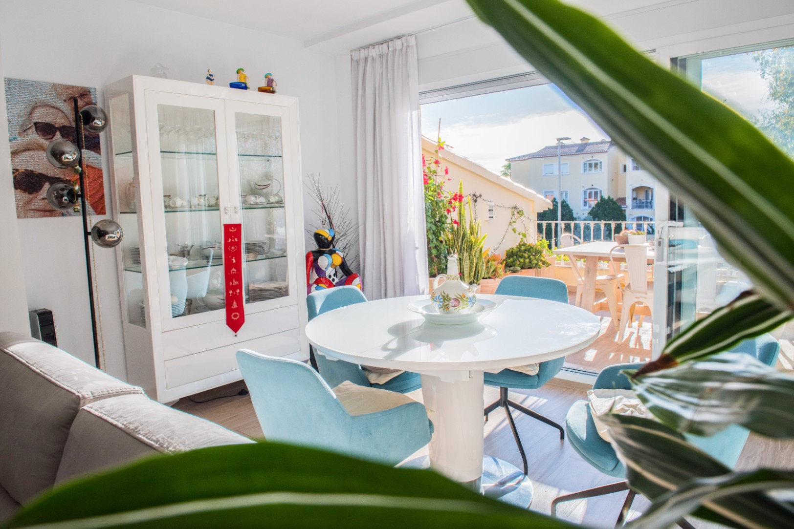 Lichte eethoek met een ronde witte tafel, blauwe gestoffeerde stoelen en een vitrinekast, grenzend aan een zonnig terras.