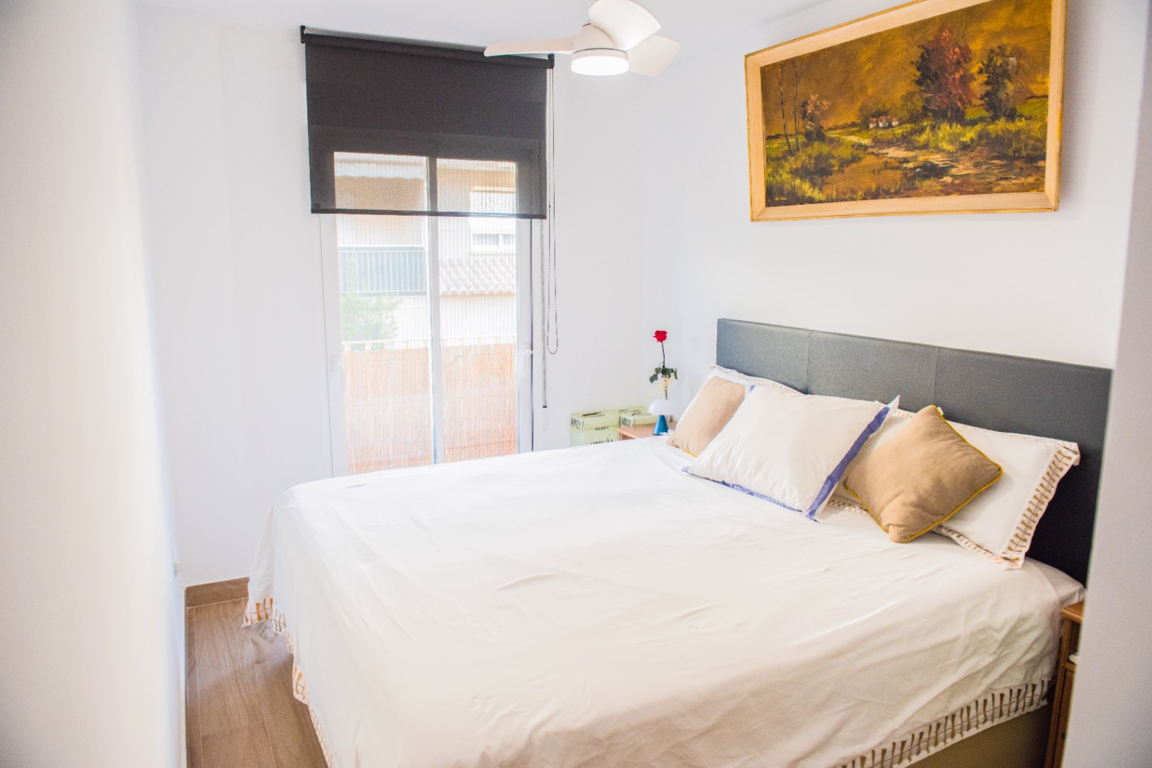 Lichte slaapkamer met grijs hoofdbord, plafondventilator en zwart rolgordijn. Een olieverfschilderij van een landschap hangt boven het bed.