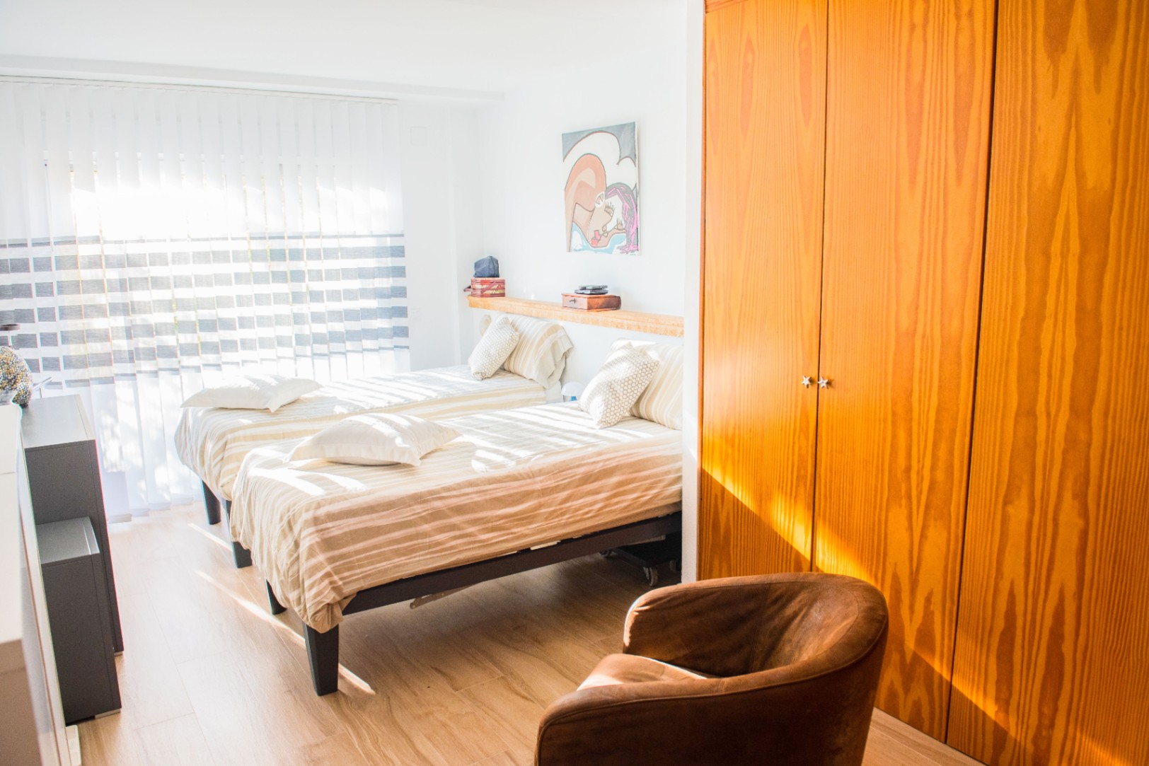 Lichte slaapkamer met twee eenpersoonsbedden, een grote houten inbouwkast en een leren fauteuil op een lichte houten vloer.
