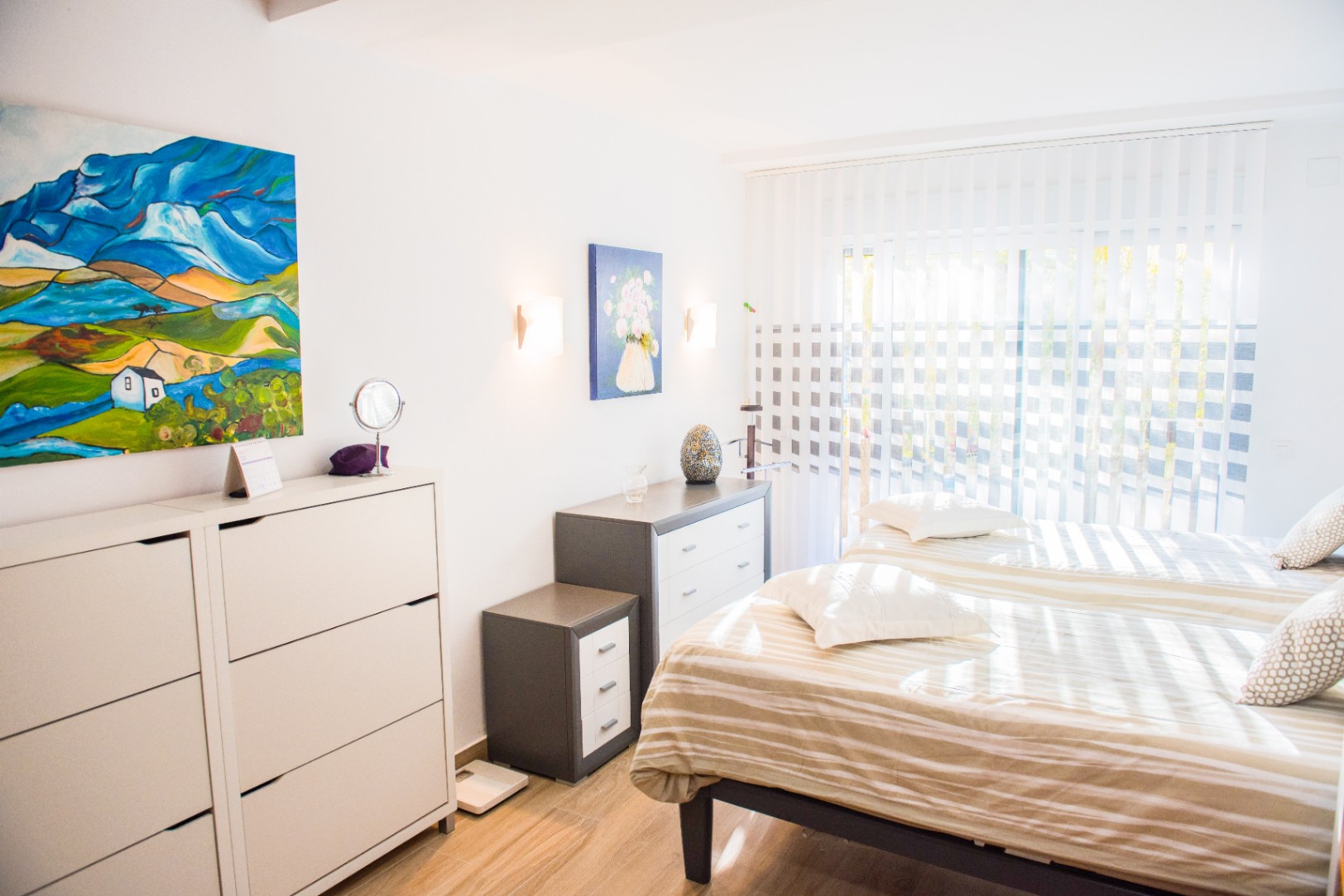 Lichte slaapkamer met twee eenpersoonsbedden, minimalistische ladekasten en kleurrijke kunst. Natuurlijk licht valt door verticale lamellen op de houtlook vloer.