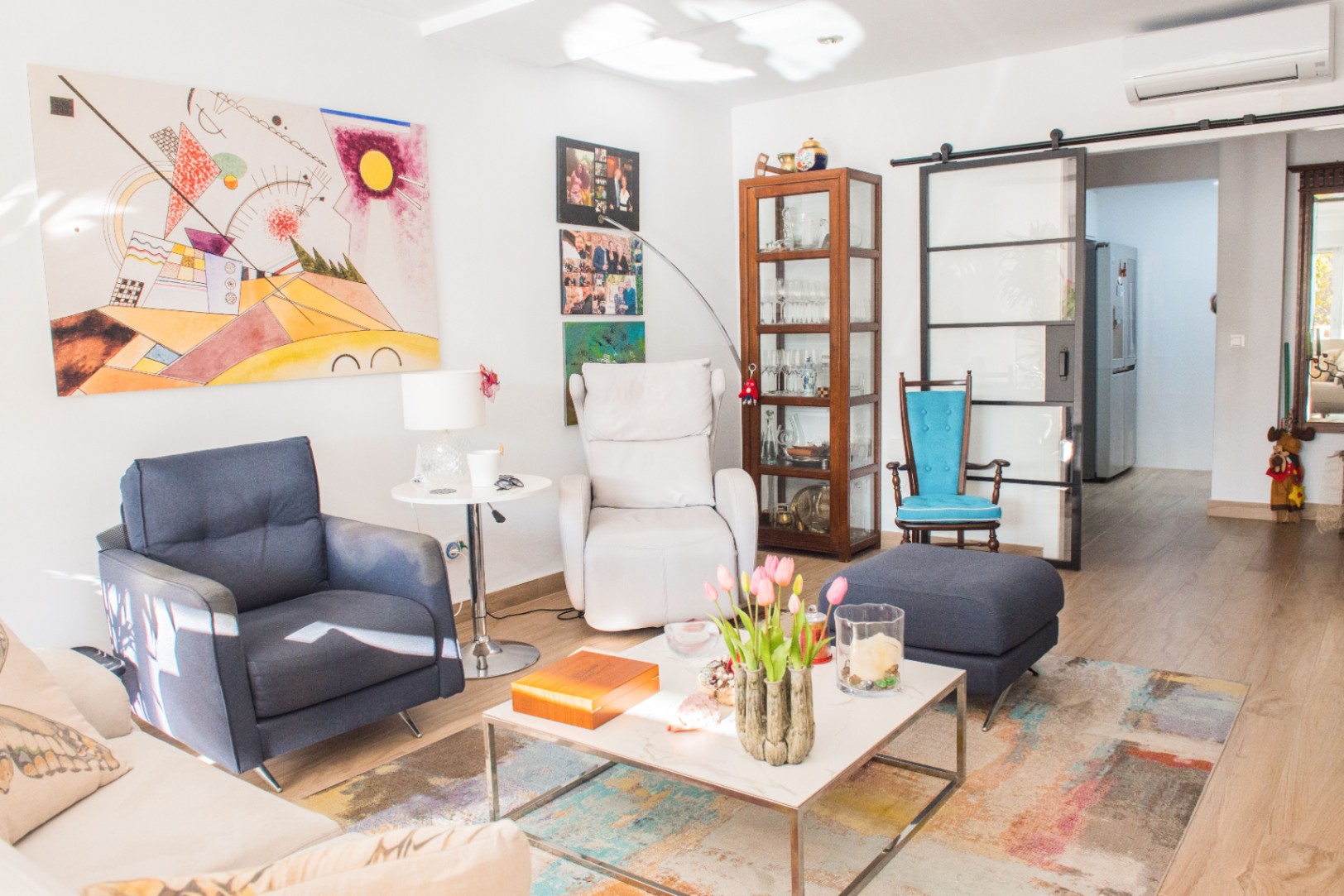 Lichte woonkamer met blauwe fauteuil, witte relaxstoel en abstracte kunst. Een glazen schuifdeur met zwart frame scheidt de ruimte van de gang.