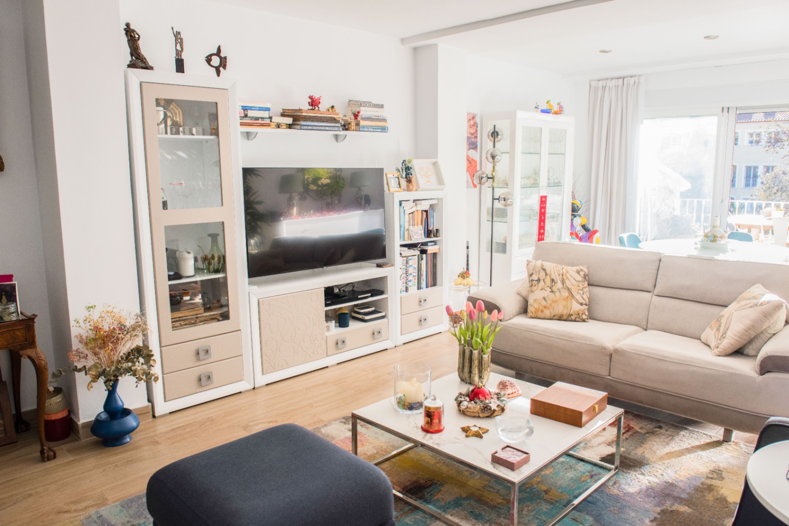Lichte woonkamer met een beige stoffen bank, marmeren salontafel en wit modulair wandmeubel. Grote glazen deuren bieden veel natuurlijk daglicht in de ruimte.