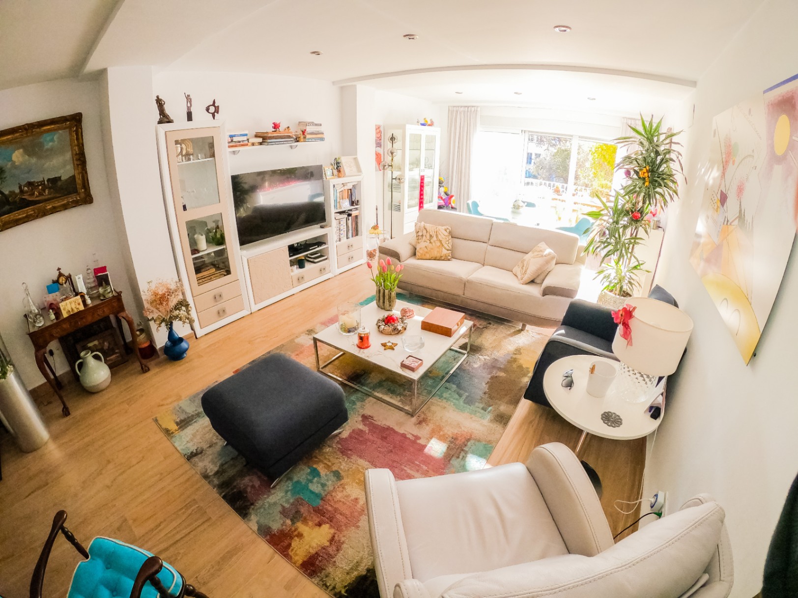 Lichte woonkamer met een kleurrijk abstract tapijt, lichte houten vloeren en een beige bank. Grote glazen schuifdeuren zorgen voor veel natuurlijk licht en toegang tot een balkon.