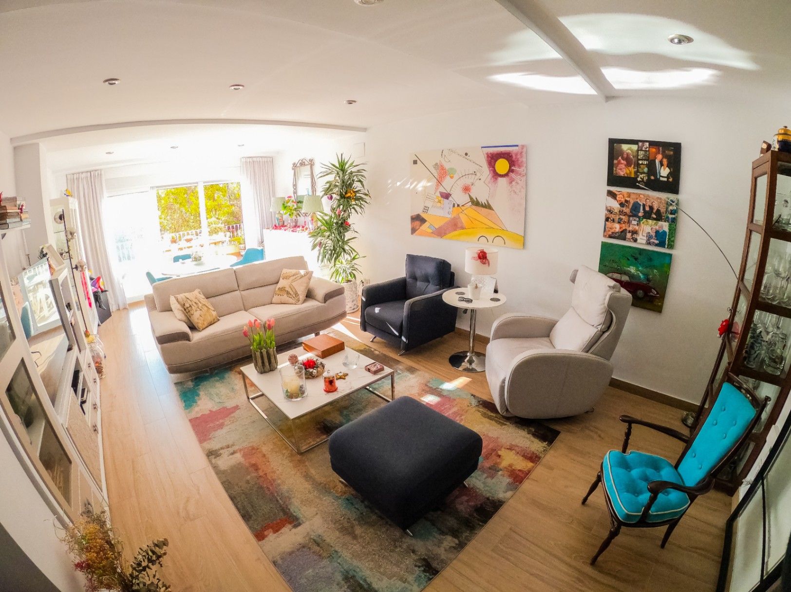 Lichte woonkamer met een lichte houten vloer, kleurrijk abstract tapijt en divers meubilair. Grote glazen schuifdeuren bieden natuurlijk licht en toegang tot een groen terras.