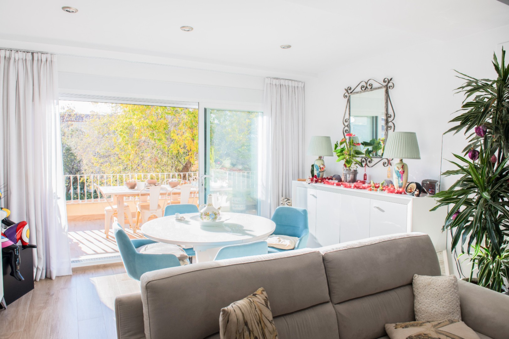 Lichte woonkamer met een ronde witte eettafel en blauwe stoelen, grenzend aan een terras met buitenmeubilair en uitzicht op de groene omgeving.