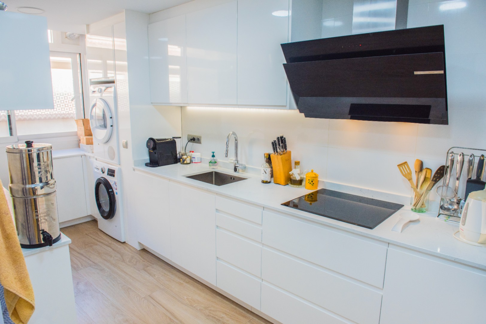 Minimalistische witte keuken met hoogglans kasten, inductiekookplaat en een geïntegreerde wasruimte met een gestapelde wasmachine en droger.
