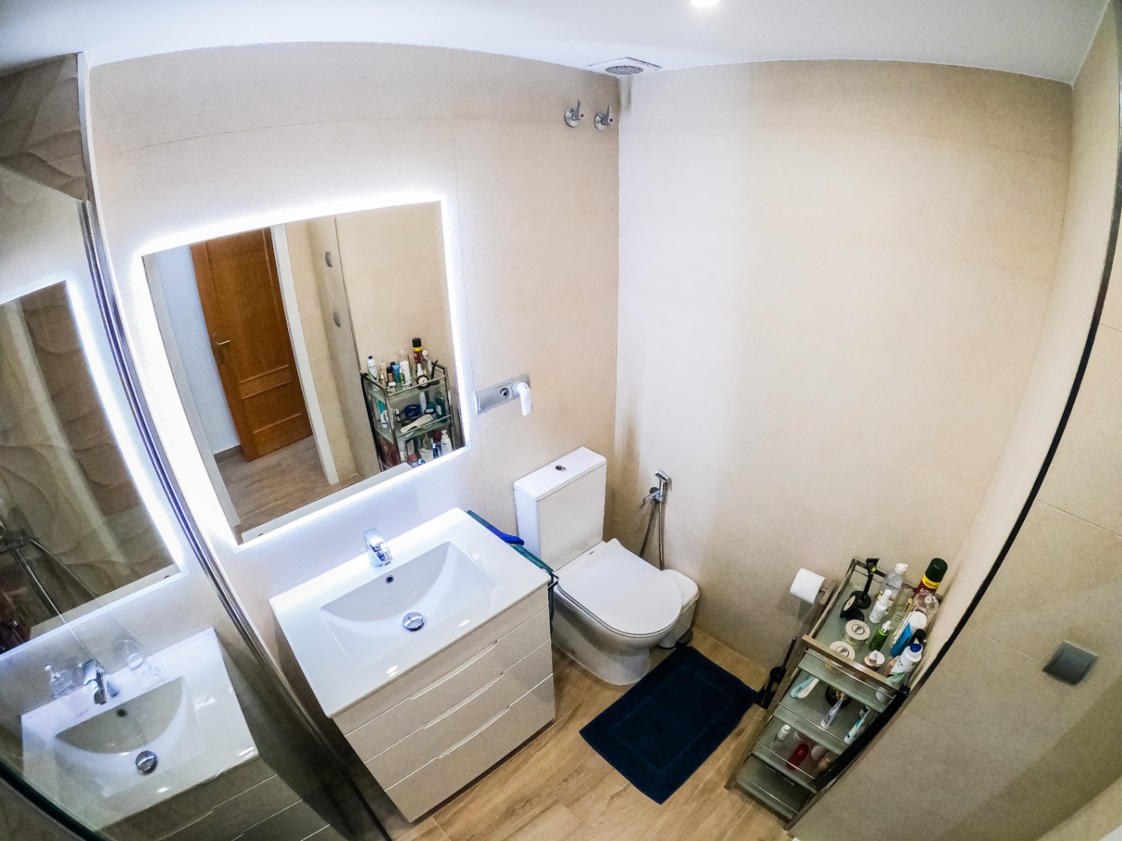 Moderne badkamer met wit wastafelmeubel, een verlichte LED-spiegel en een vloer met houtlook. De ruimte is afgewerkt met beige wandtegels en een staand toilet.