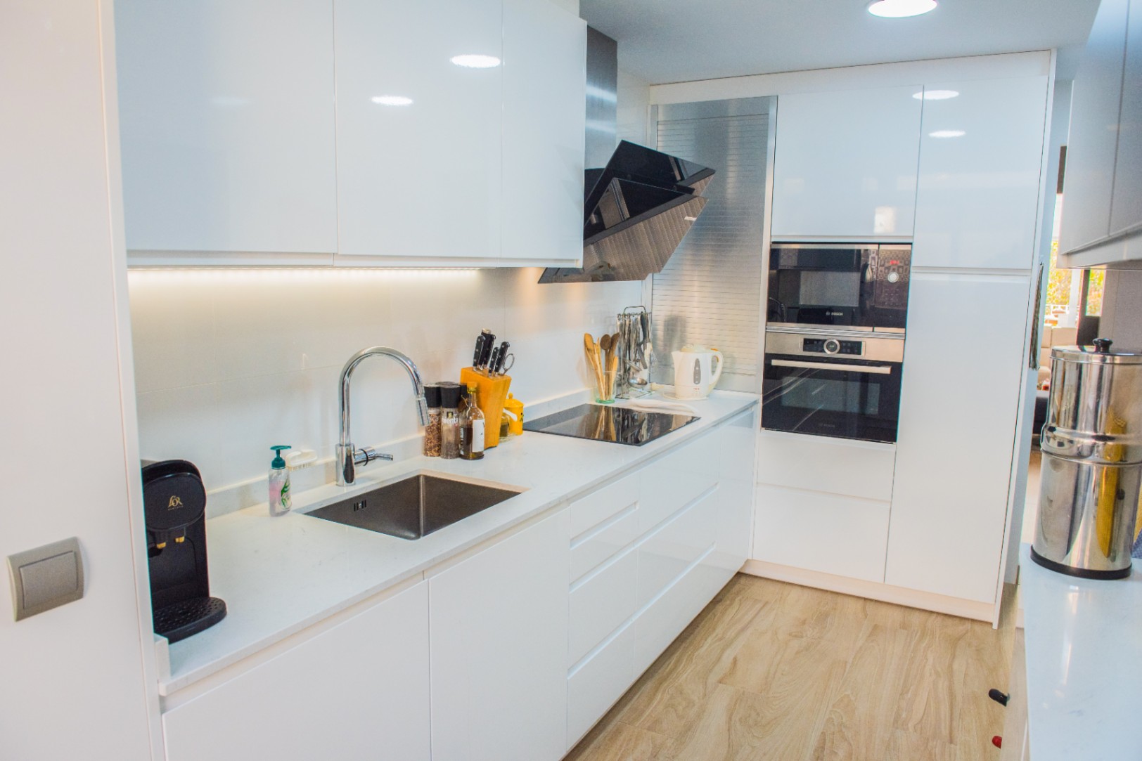 Moderne keuken met witte hoogglanskasten, geïntegreerde Bosch-apparatuur en een inductiekookplaat. Voorzien van een wit kwarts werkblad en een lichte houten vloer.