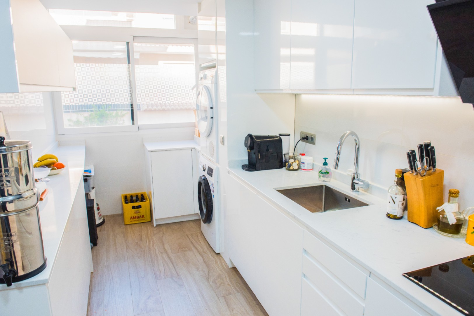 Moderne parallelkeuken met greeploze witte kasten, geïntegreerde wasruimte en wit werkblad. Voorzien van LED-onderbouwverlichting en een lichte houtlook vloer.