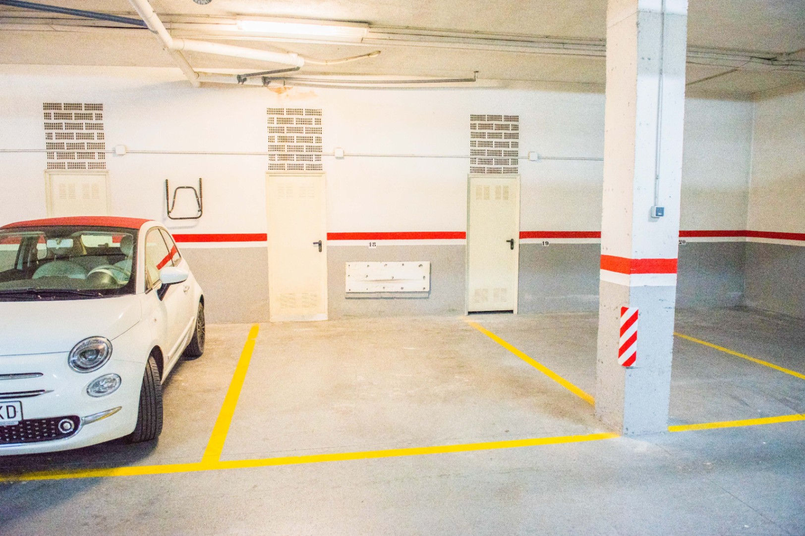 Ondergrondse parkeerplaats met genummerde vakken, gele vloerlijnen en wit-grijze wanden met rode accenten en zichtbare plafondleidingen.