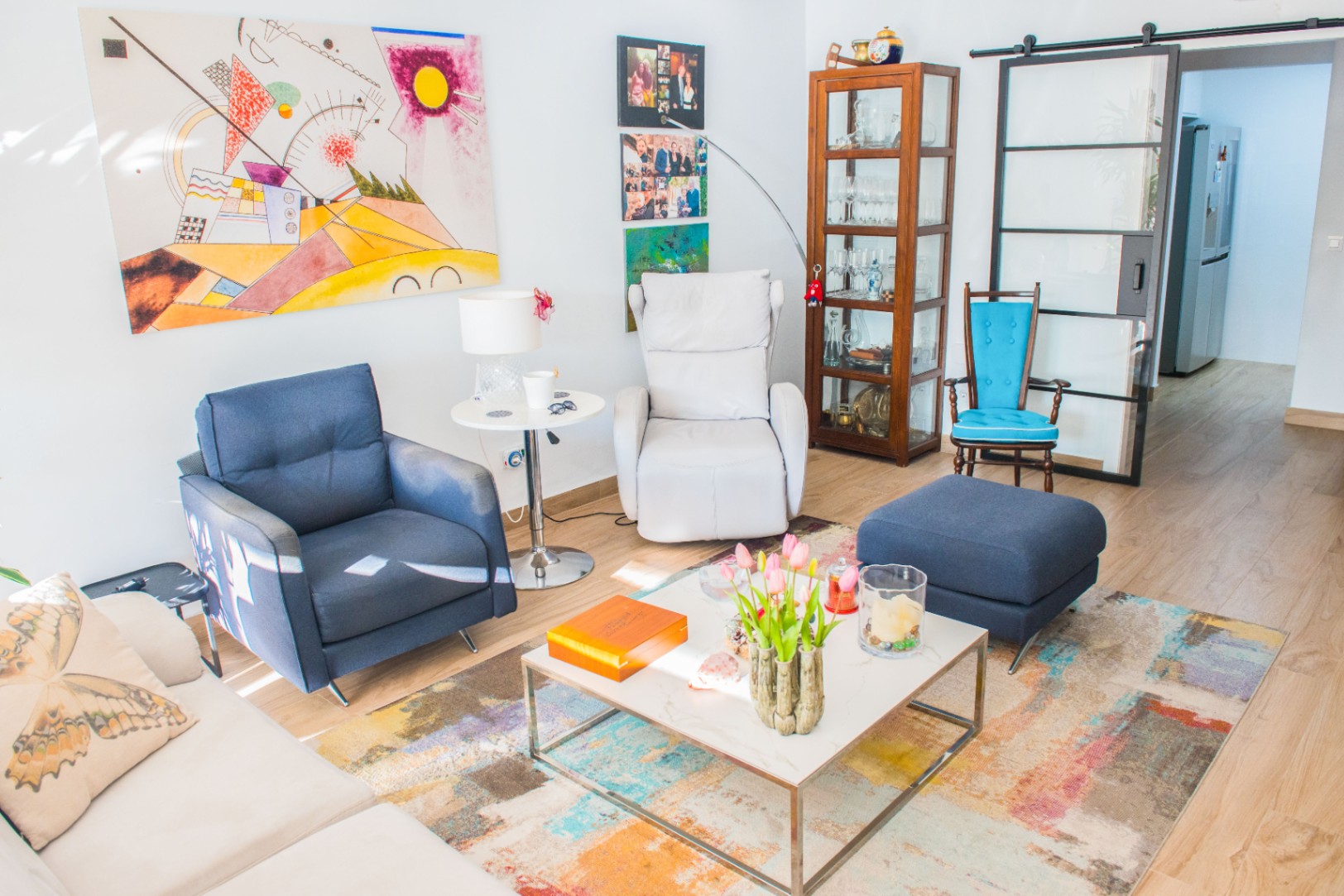 Woonkamer met blauwe fauteuil, witte relaxstoel en houten vitrinekast. Voorzien van abstracte kunst, een kleurrijk tapijt en een glazen schuifdeur met zwart industrieel frame.