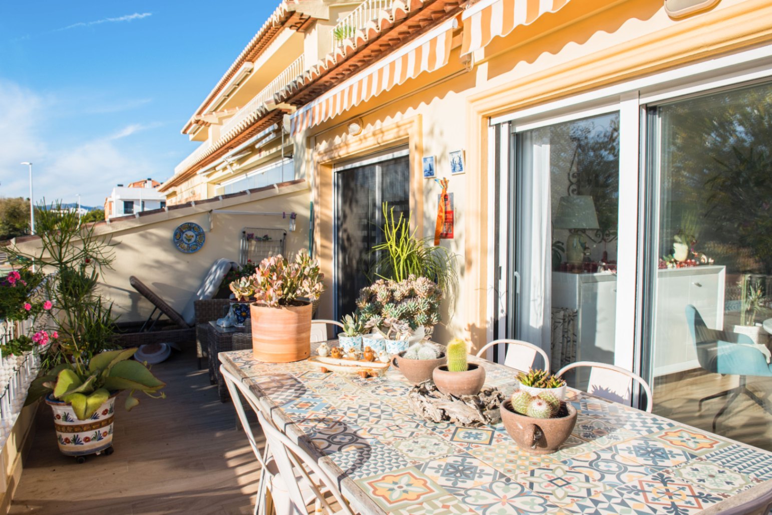 Zonnig mediterraan terras met een betegelde mozaïektafel, vetplanten in potten en grote glazen schuifdeuren onder een gestreepte zonnewering.