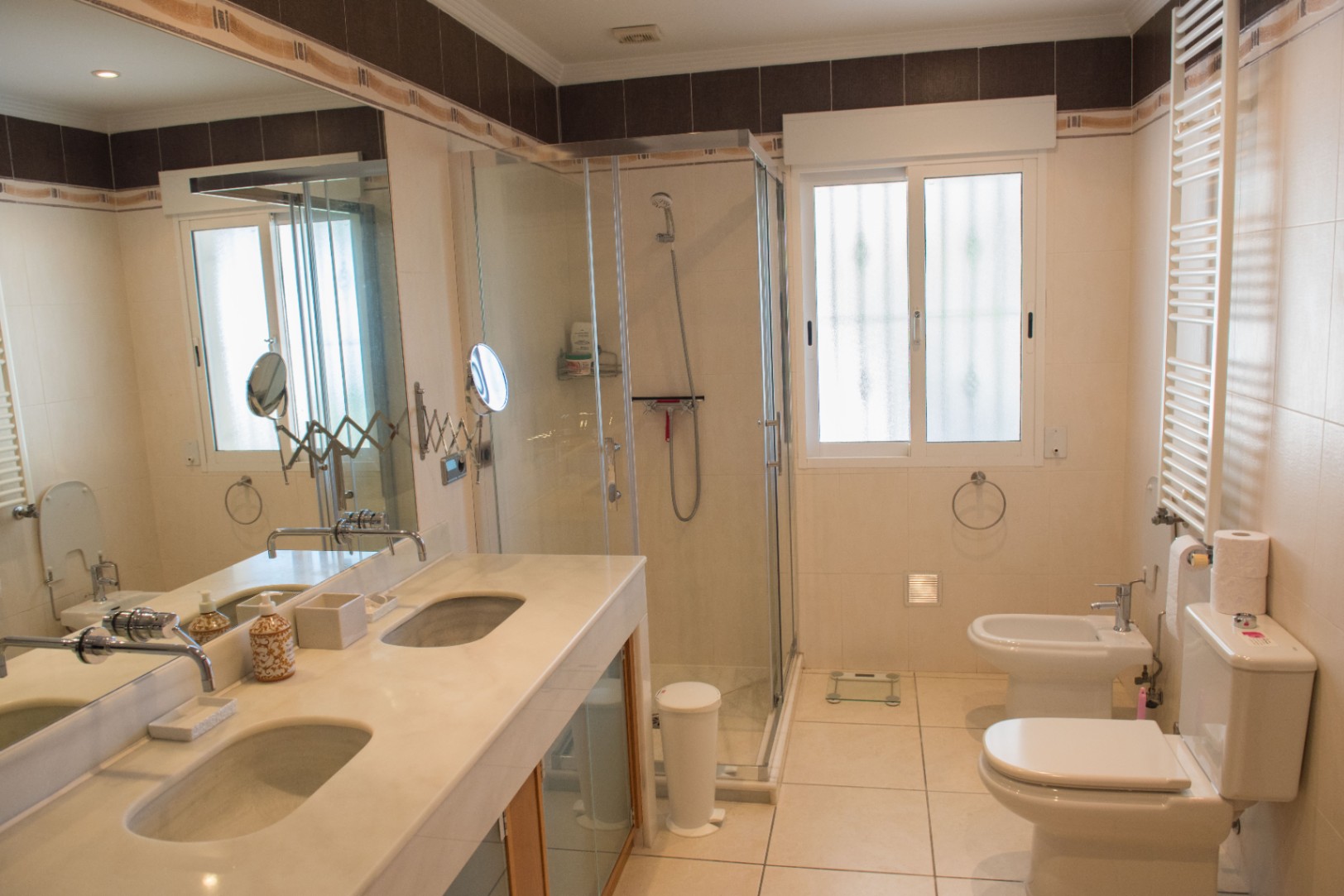 Badkamer met dubbele marmeren wastafel, een glazen douchecabine en wit sanitair, afgewerkt met neutrale tegels en een grote wandspiegel.
