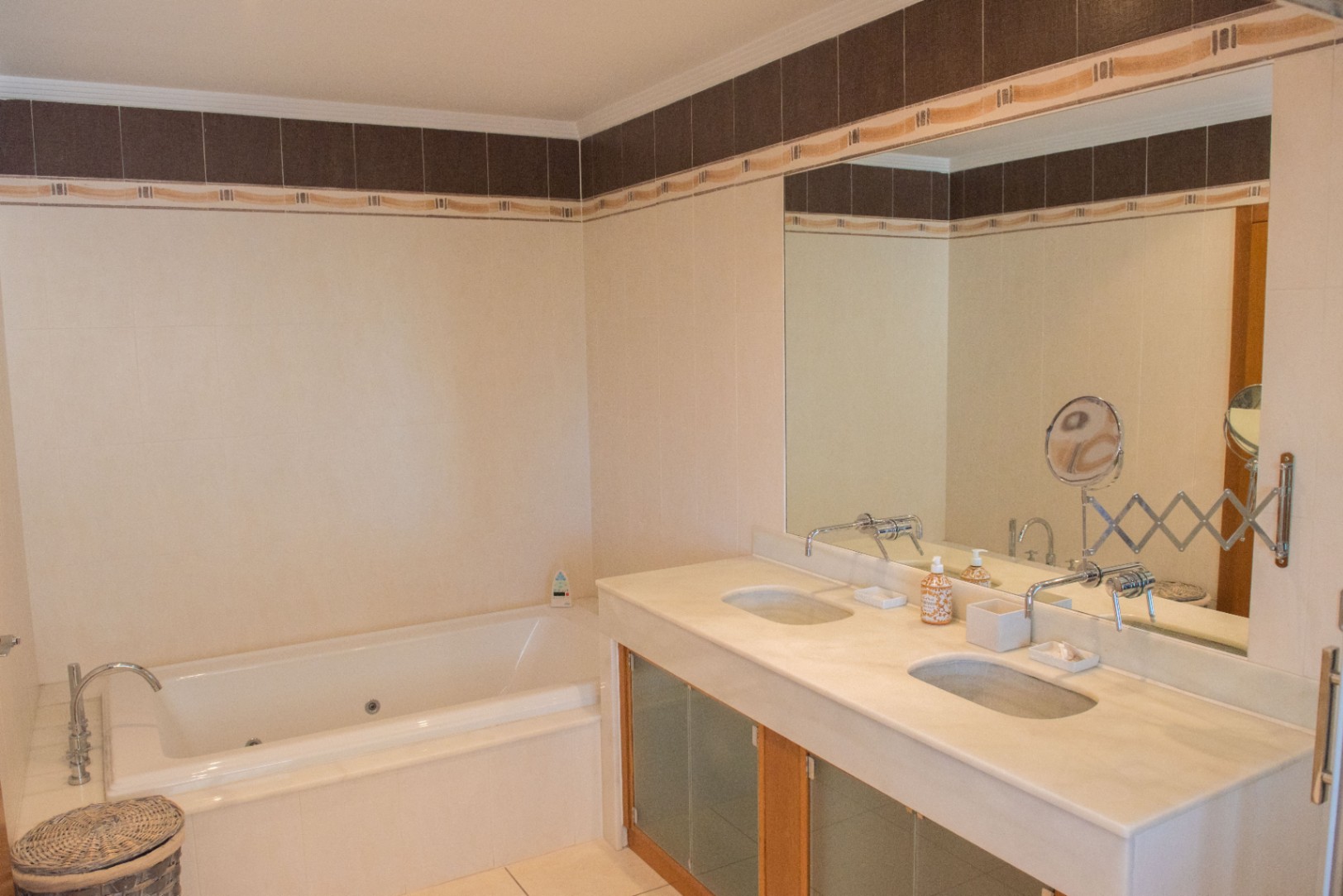 Badkamer met dubbele wastafel op een stenen blad, wandkranen en een ingebouwd bubbelbad, voorzien van wandtegels met een decoratieve sierlijst.