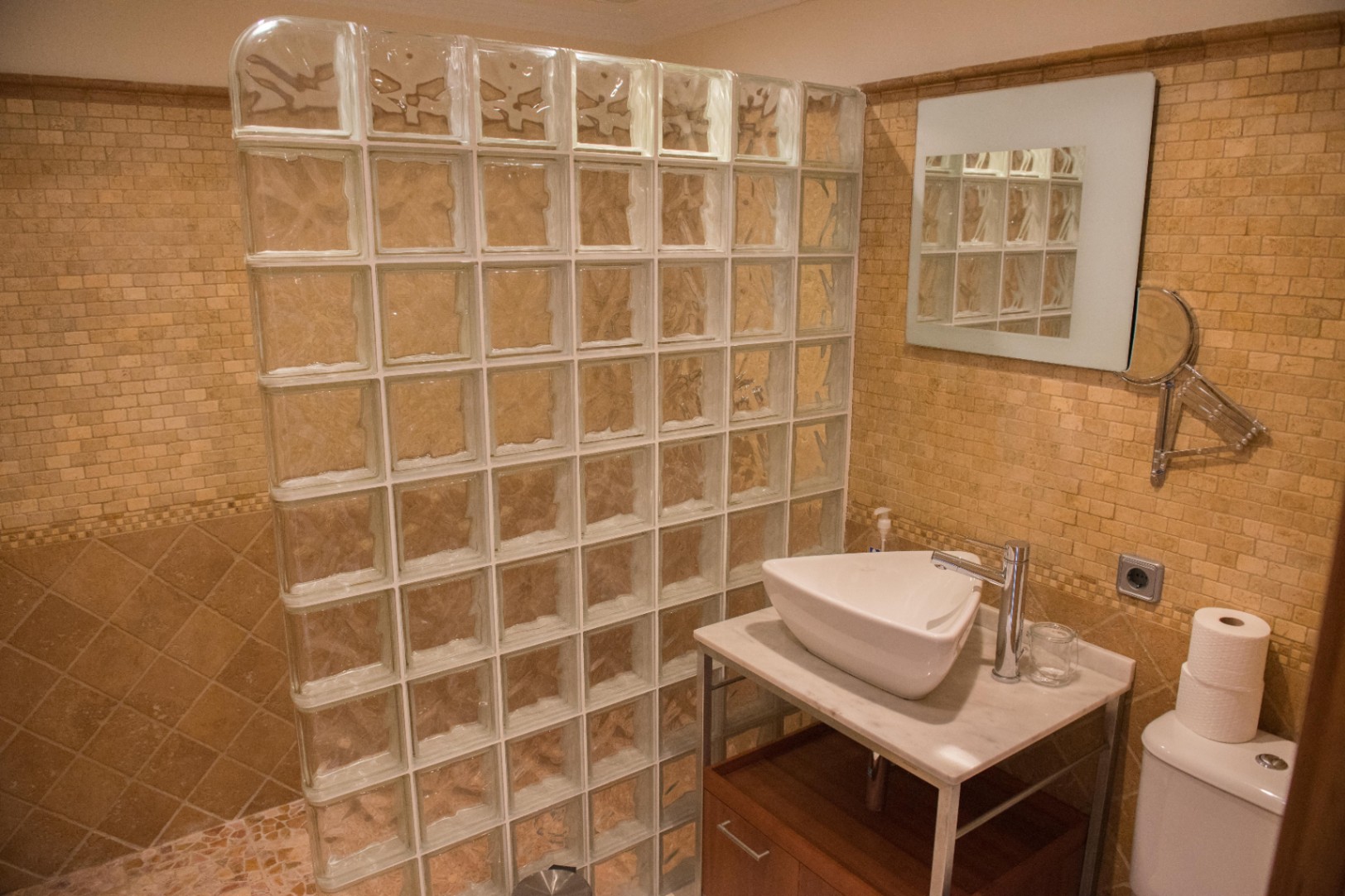 Badkamer met een douchewand van glasbouwstenen, beige mozaïektegels en een opzetwastafel op een marmeren blad met houten onderkast.