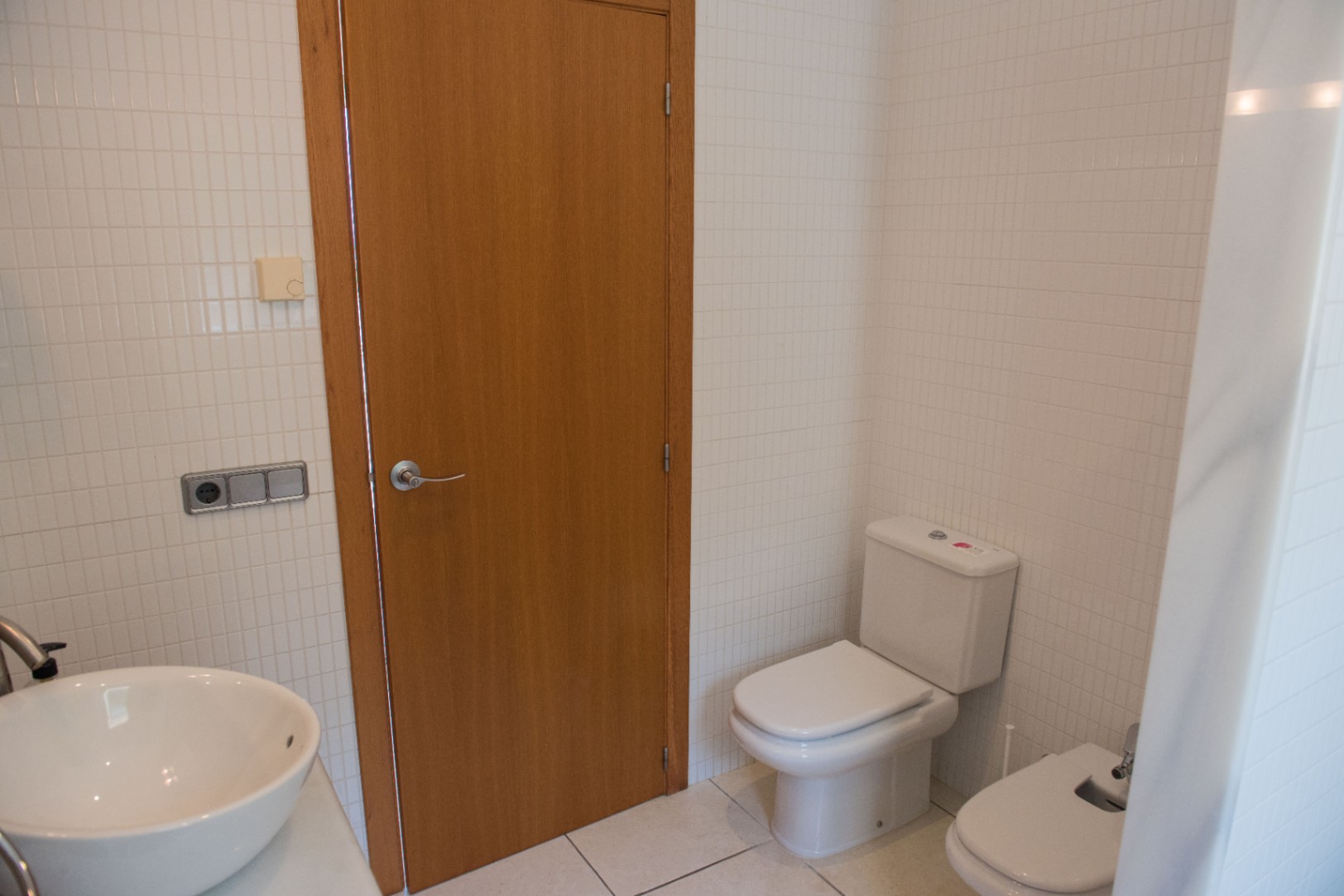 Badkamer met witte vierkante wandtegels, houten deur, keramisch toilet, bidet en een witte opzetwastafel met chromen kranen.