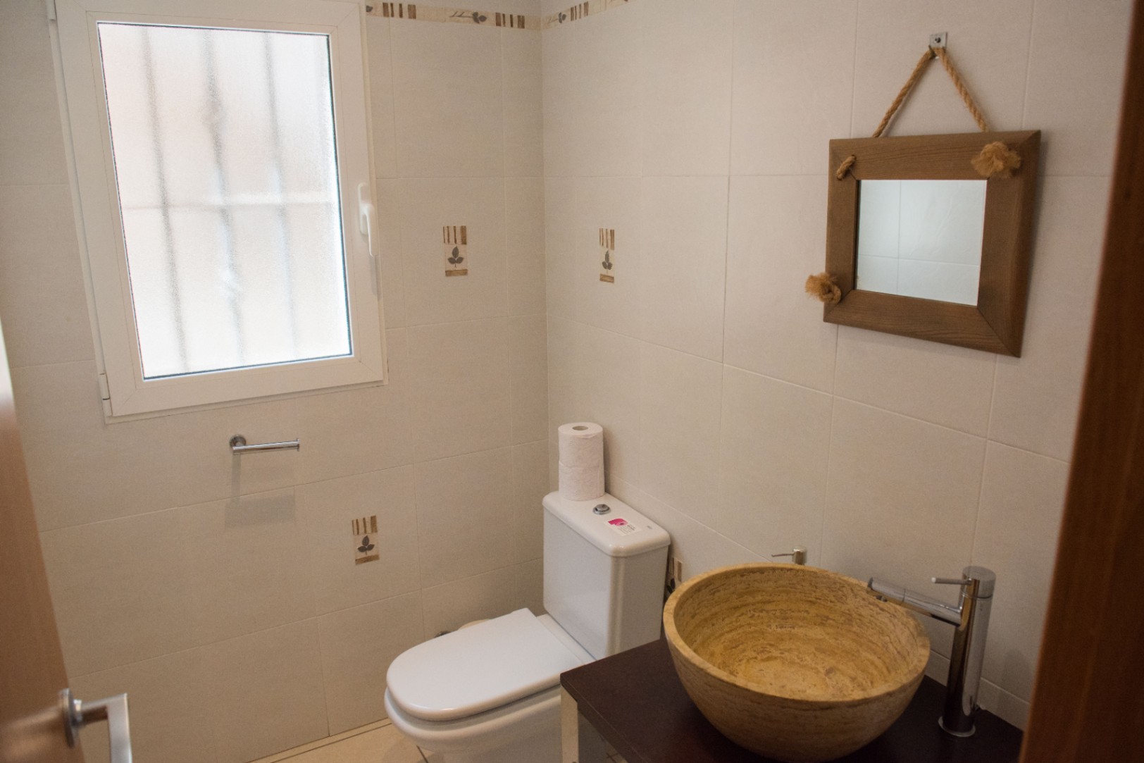 Compact toilet met een natuurstenen waskom op een donker houten meubel, een rustieke spiegel met touwophanging en licht betegelde wanden met matglas.