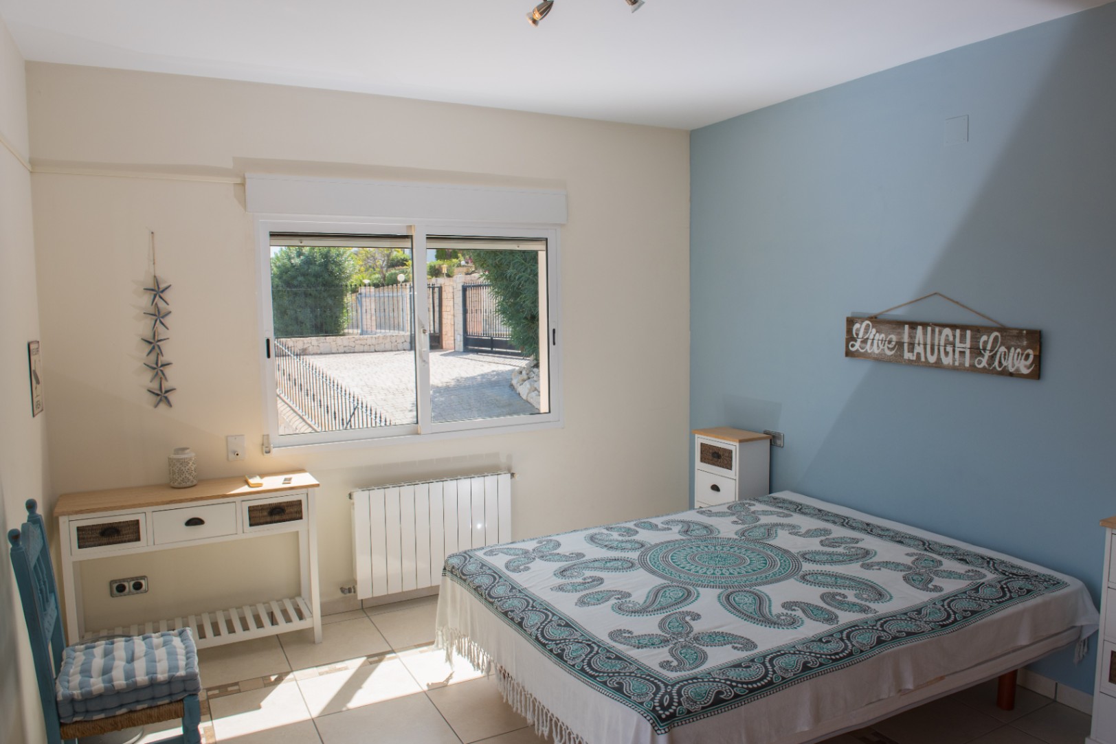 Lichte slaapkamer met blauwe accentmuur, tegelvloer en raam met uitzicht op de omheinde oprit. Inclusief witte consoletafel en wandradiator.