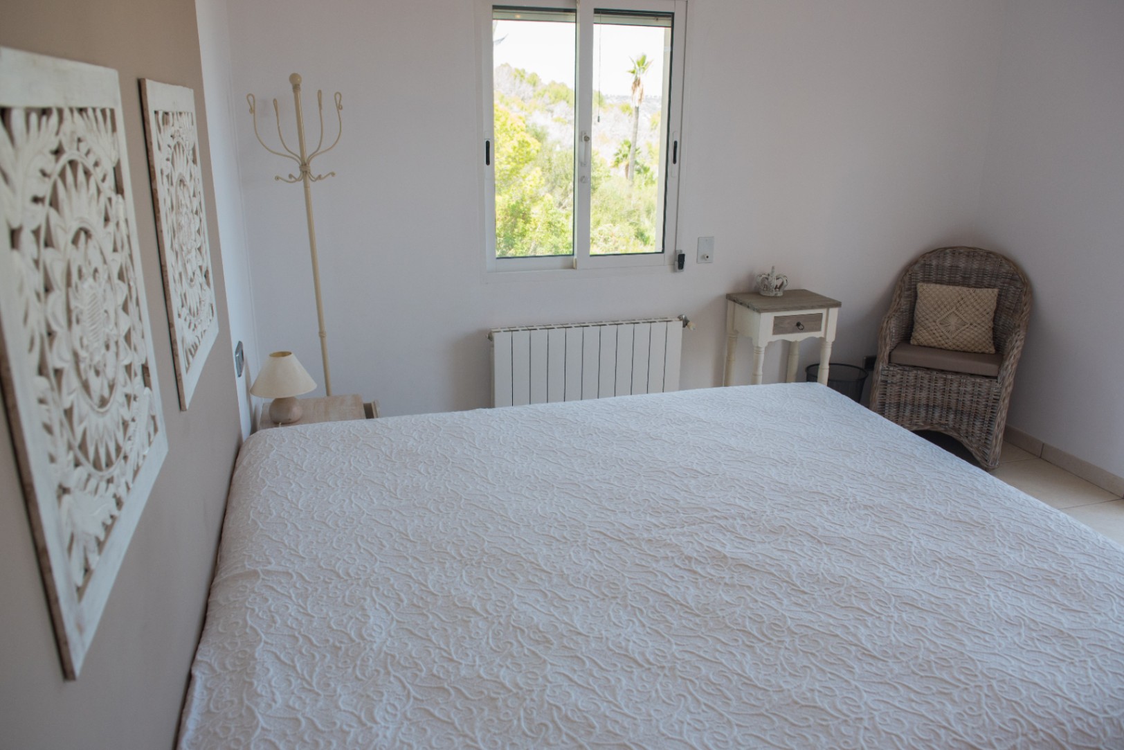 Lichte slaapkamer met een witte sprei met textuur, gesneden wanddecoratie en een rieten fauteuil. Het raam biedt uitzicht op de omliggende groene beplanting.