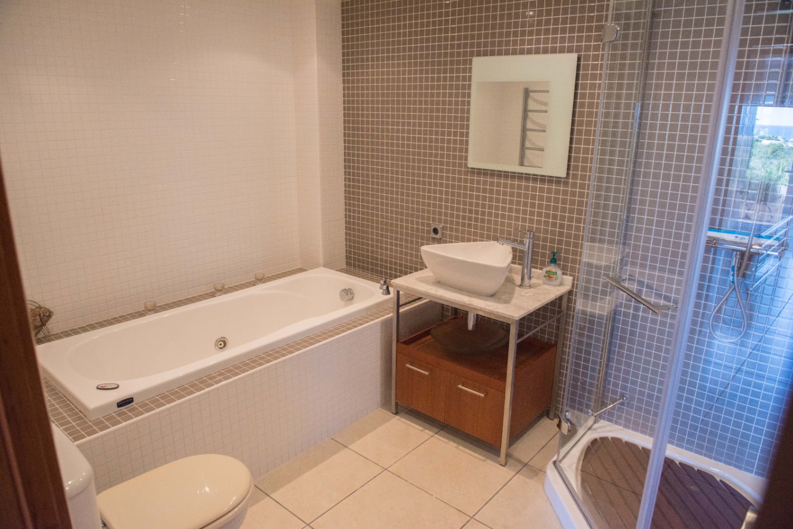 Moderne badkamer met ingebouwd ligbad, glazen douchecabine met houten vlonder en een opzetwastafel op een marmeren blad met taupe mozaïektegels.