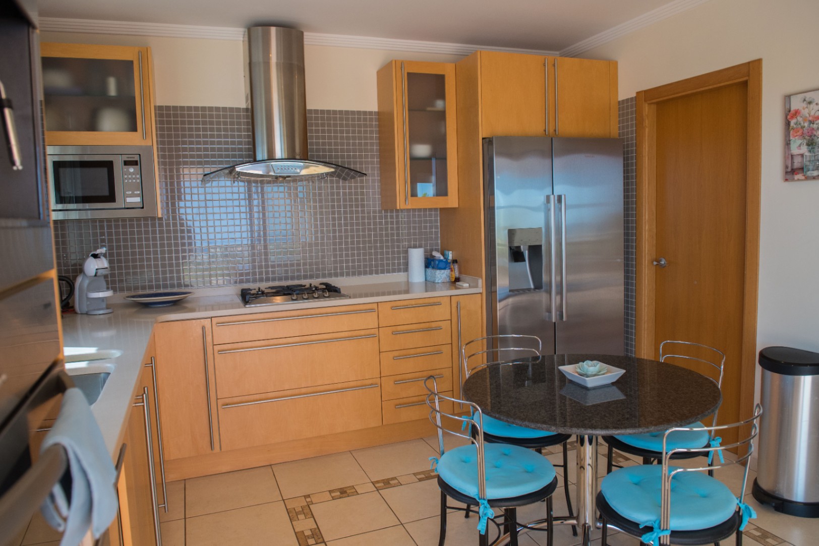 Moderne keuken met lichte houten kasten, een grijze mozaïek achterwand en roestvrijstalen apparatuur. Voorzien van een ronde granieten eettafel met blauwe kussens.