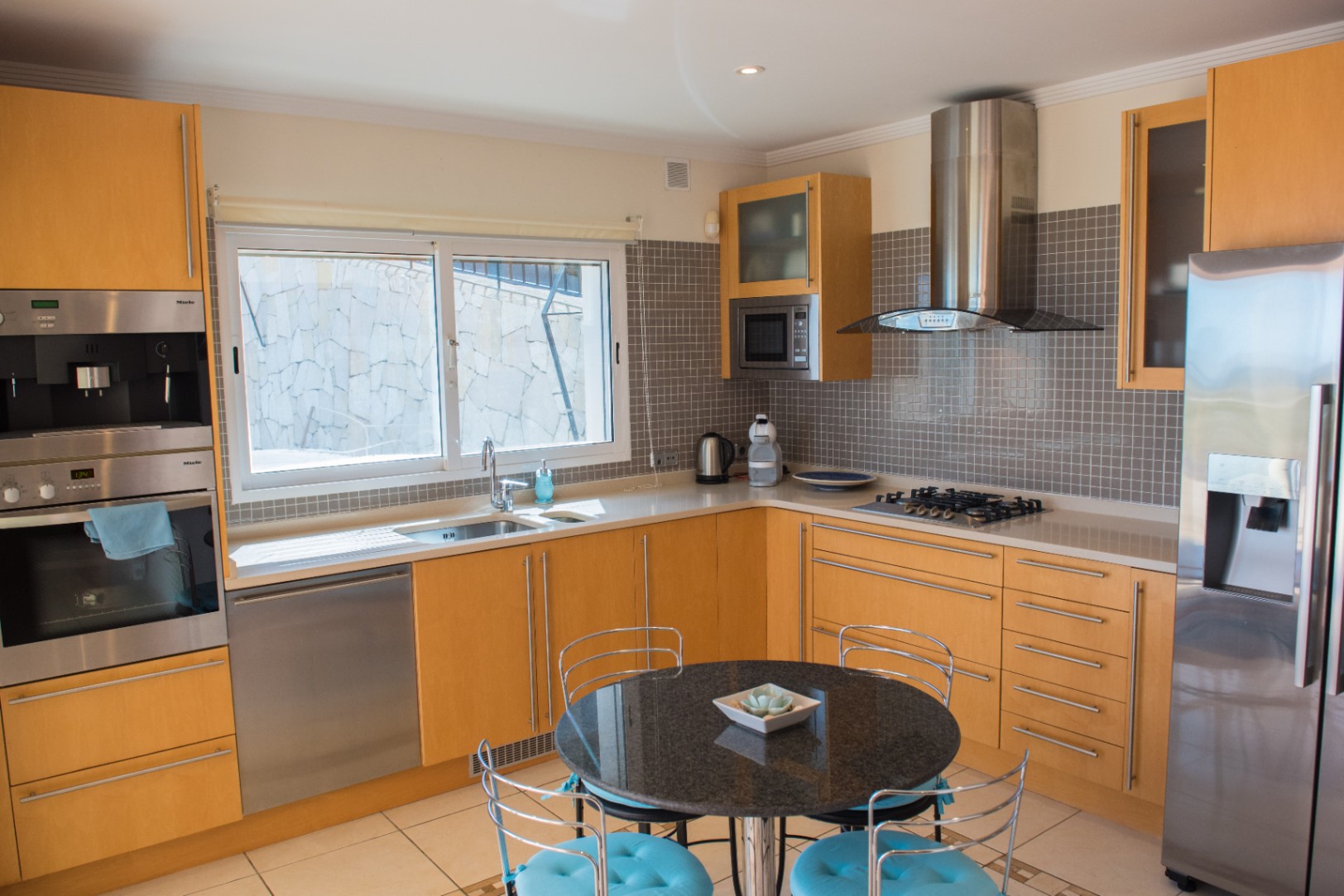 Moderne keuken met lichte houten kasten, roestvrijstalen Miele-apparatuur en een grijze mozaïek achterwand. Voorzien van een zwarte granieten tafel en wit aanrechtblad.