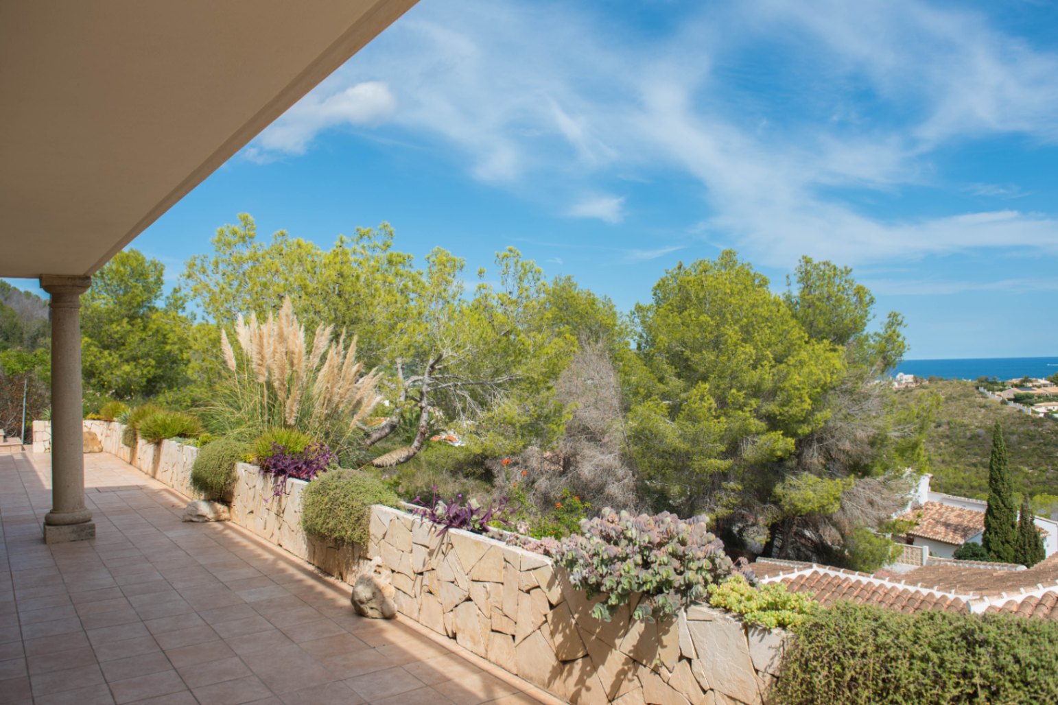 Overdekt terras met tegelvloer en een natuurstenen plantenbak met mediterrane beplanting, met uitzicht op pijnbomen en de zee.