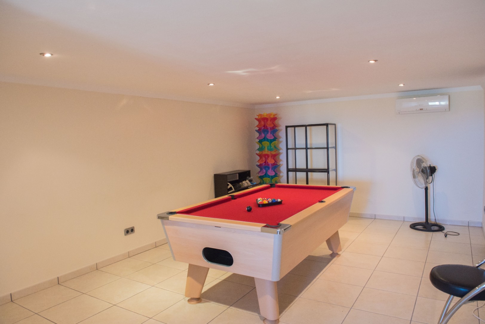 Ruime ontspanningsruimte met een biljarttafel met rood laken, lichte tegelvloer, inbouwspots en wandgemonteerde airconditioning.