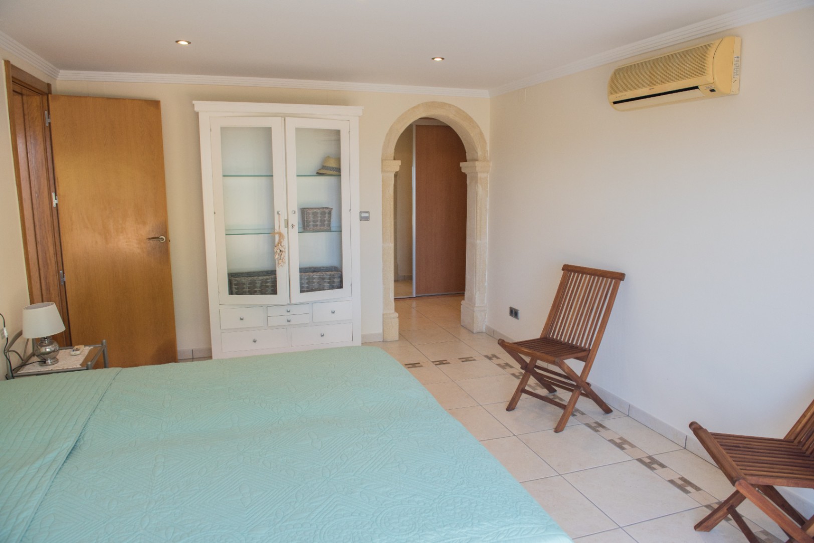 Slaapkamer met een natuurstenen boog, tegelvloer met decoratieve rand, witte vitrinekast en houten klapstoelen. Voorzien van airconditioning en inbouwspots.