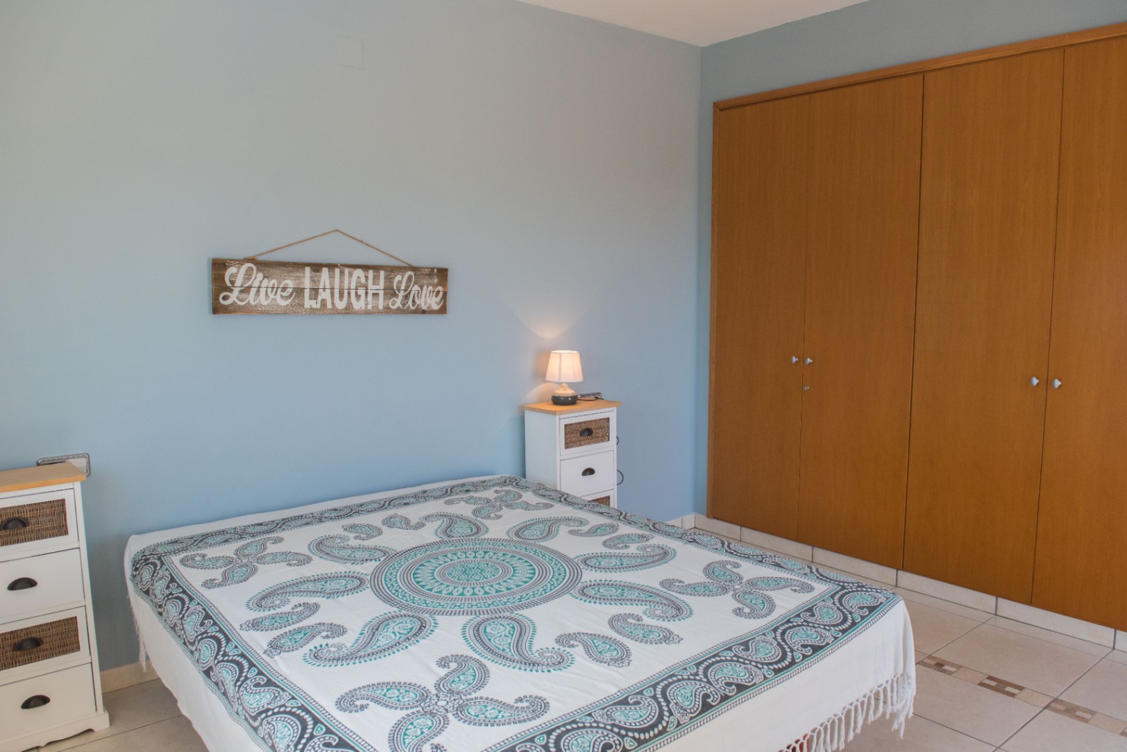 Slaapkamer met lichtblauwe wanden, een grote houten inbouwkast en witte meubels met rieten accenten. De ruimte is voorzien van een tegelvloer en een bedsprei met motief.