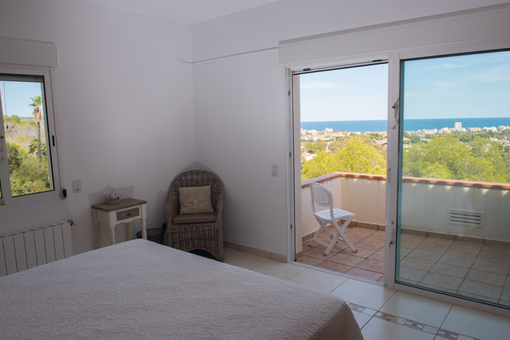 Slaapkamer met toegang tot een balkon met panoramisch uitzicht op zee. Voorzien van een witte tegelvloer, een rieten fauteuil en een glazen schuifpui.