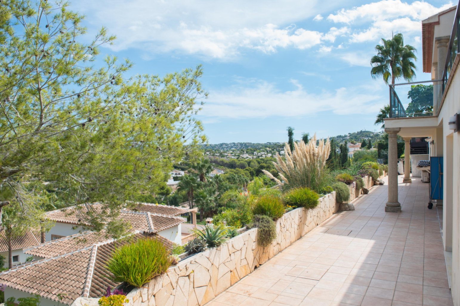 Verhoogd terras met beige tegels en een natuurstenen plantenbak. Biedt een panoramisch uitzicht over de vallei, omliggende villa's en mediterrane beplanting.