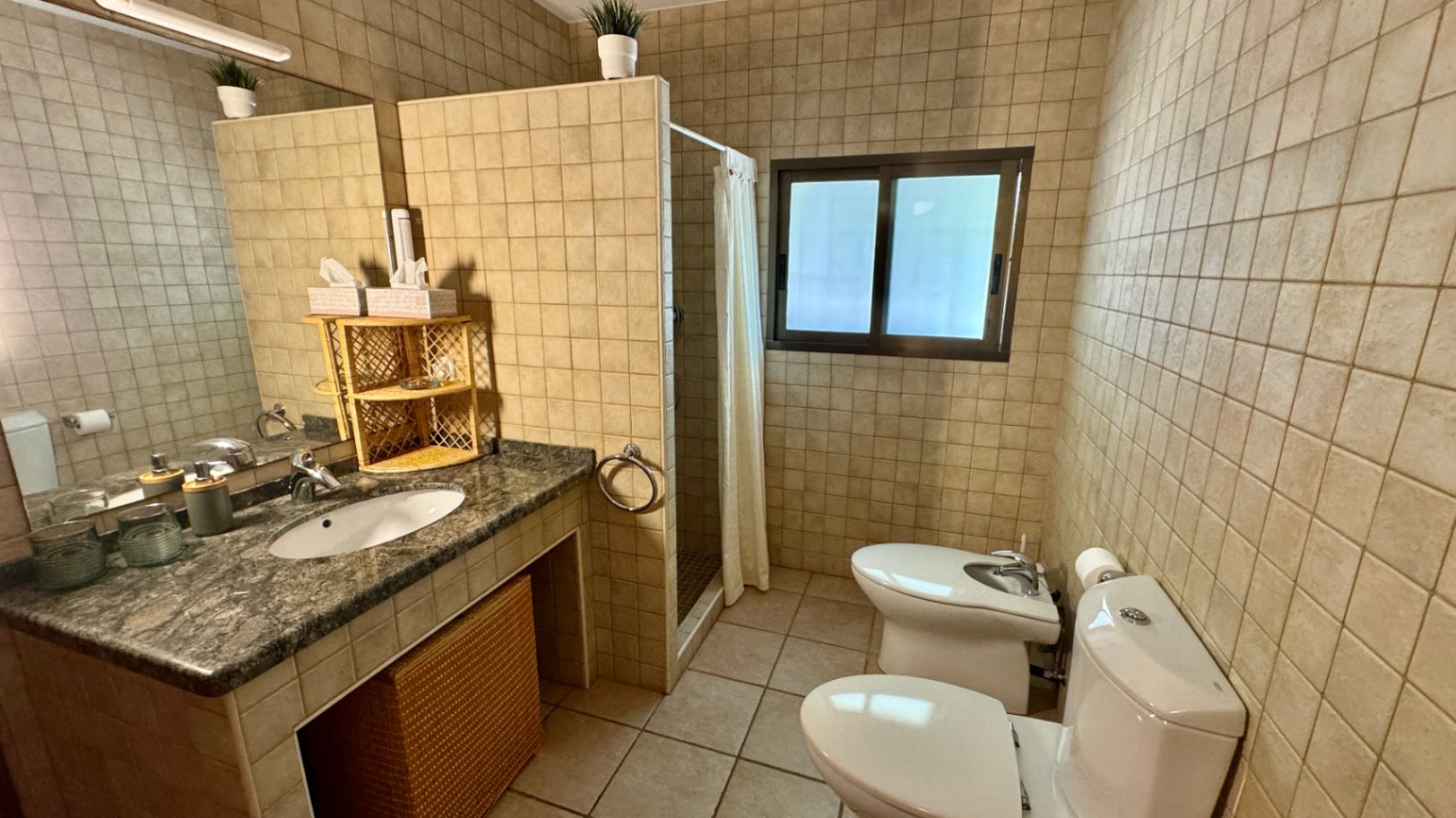 Badkamer met granieten wastafelblad, ingebouwde wasbak en rieten opbergmanden. Voorzien van beige wandtegels, inloopdouche met gordijn en een toilet met bidet.