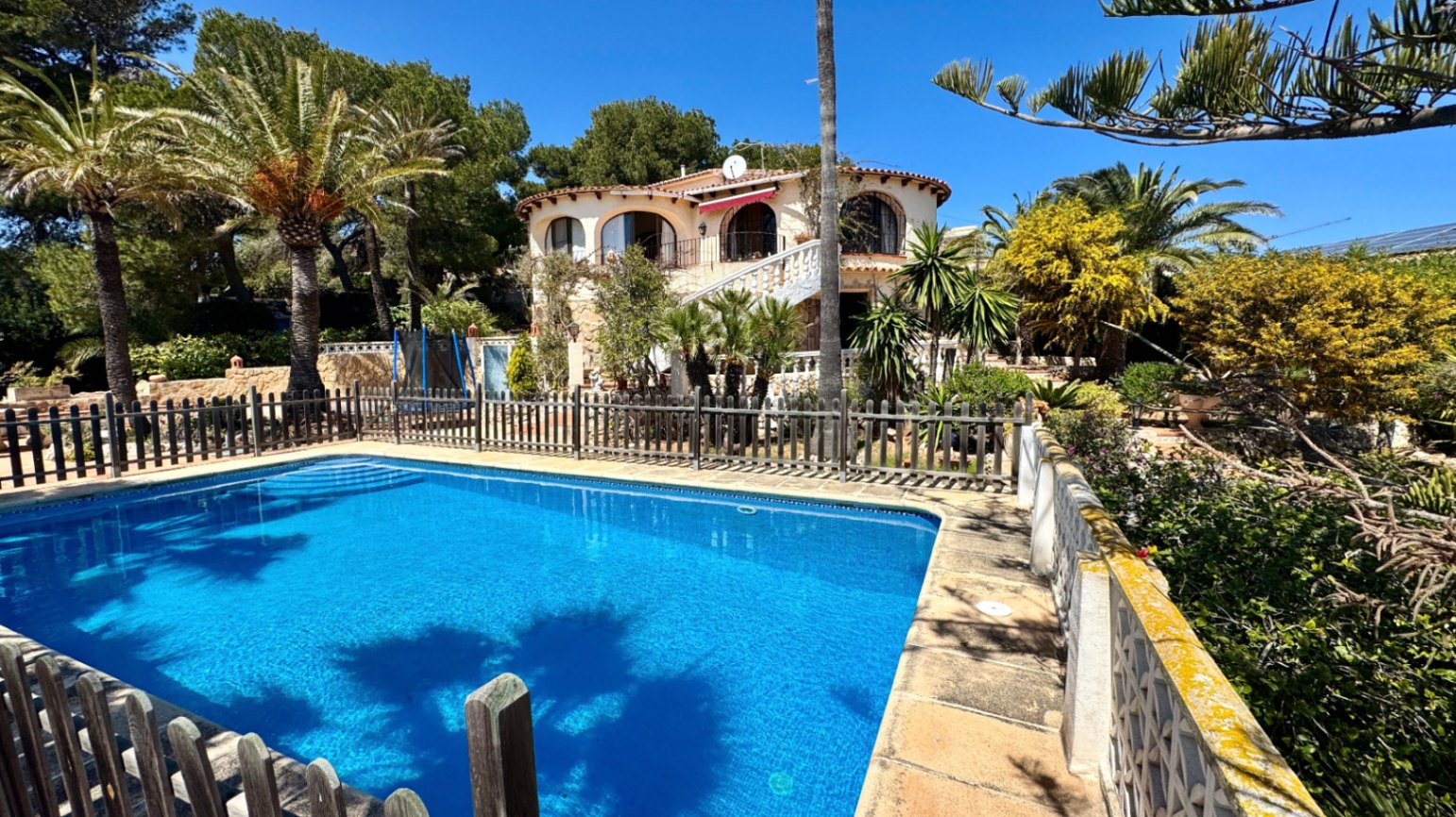 Buitenaanzicht van een omheind rechthoekig zwembad bij een mediterrane villa met boogvormige balkons, omgeven door volgroeide palmbomen.