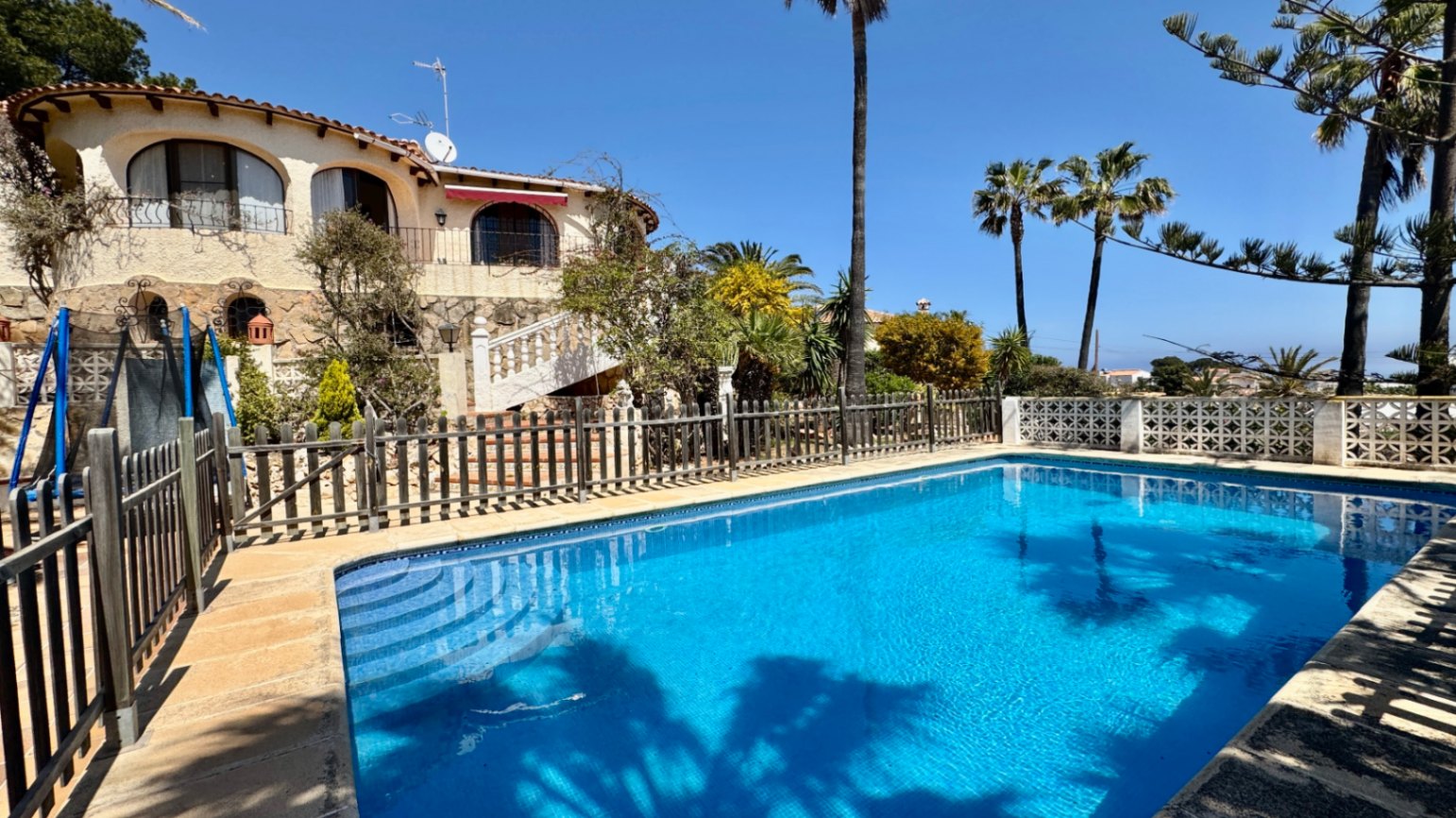 Buitenzwembad met blauwe tegels en inlooptrap, omgeven door een houten omheining en een mediterrane villa met stenen geveldetails en boogvormige balkons.