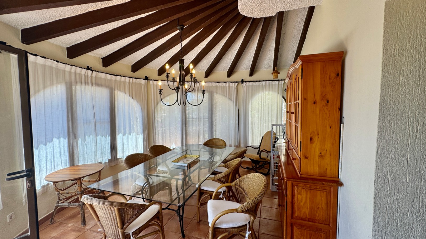 Eethoek met een radiaal houten balkenplafond, een glazen tafel met rieten stoelen en een houten buffetkast op een terracotta vloer.