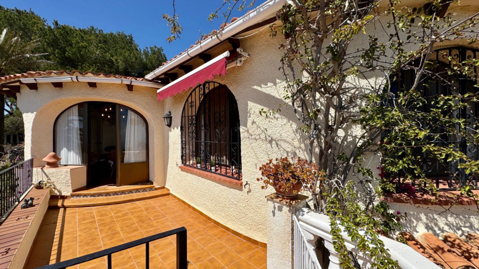 Exterieur van een mediterrane villa met een terracotta terras, boogvensters met smeedijzeren roosters en een rode uitvalscherm.