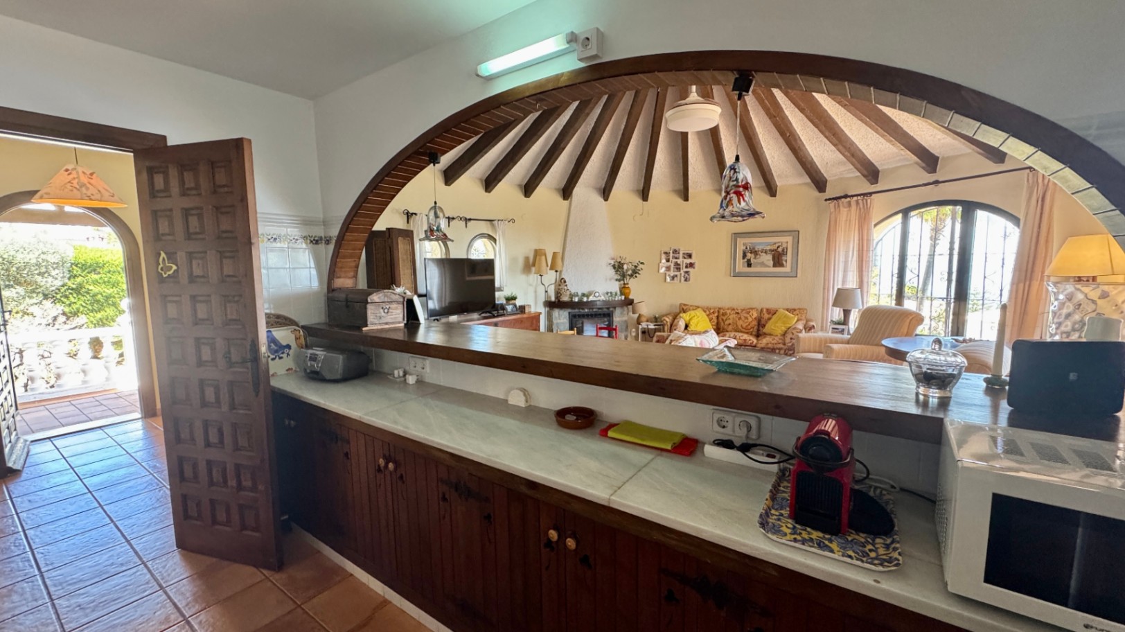 Keukenbar met marmeren aanrechtblad en houten kasten, uitkijkend door een grote houten boog op een ronde woonkamer met radiale plafondbalken.