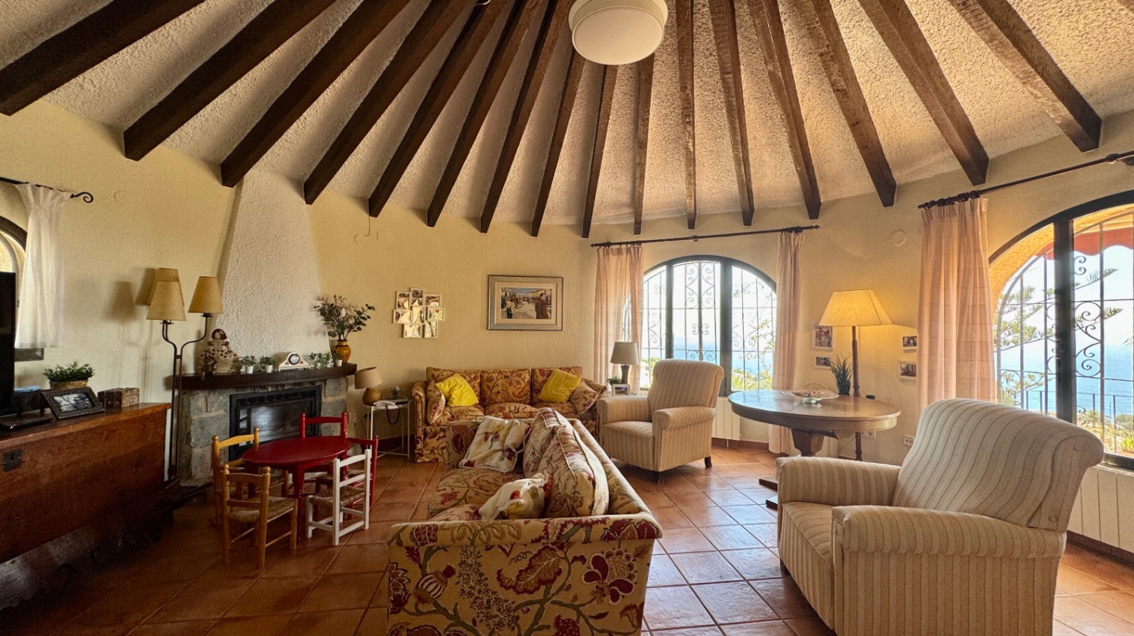 Ronde woonkamer met een kegelvormig plafond met radiale houten balken, terracotta vloer, stenen open haard en boogvensters met uitzicht op zee.