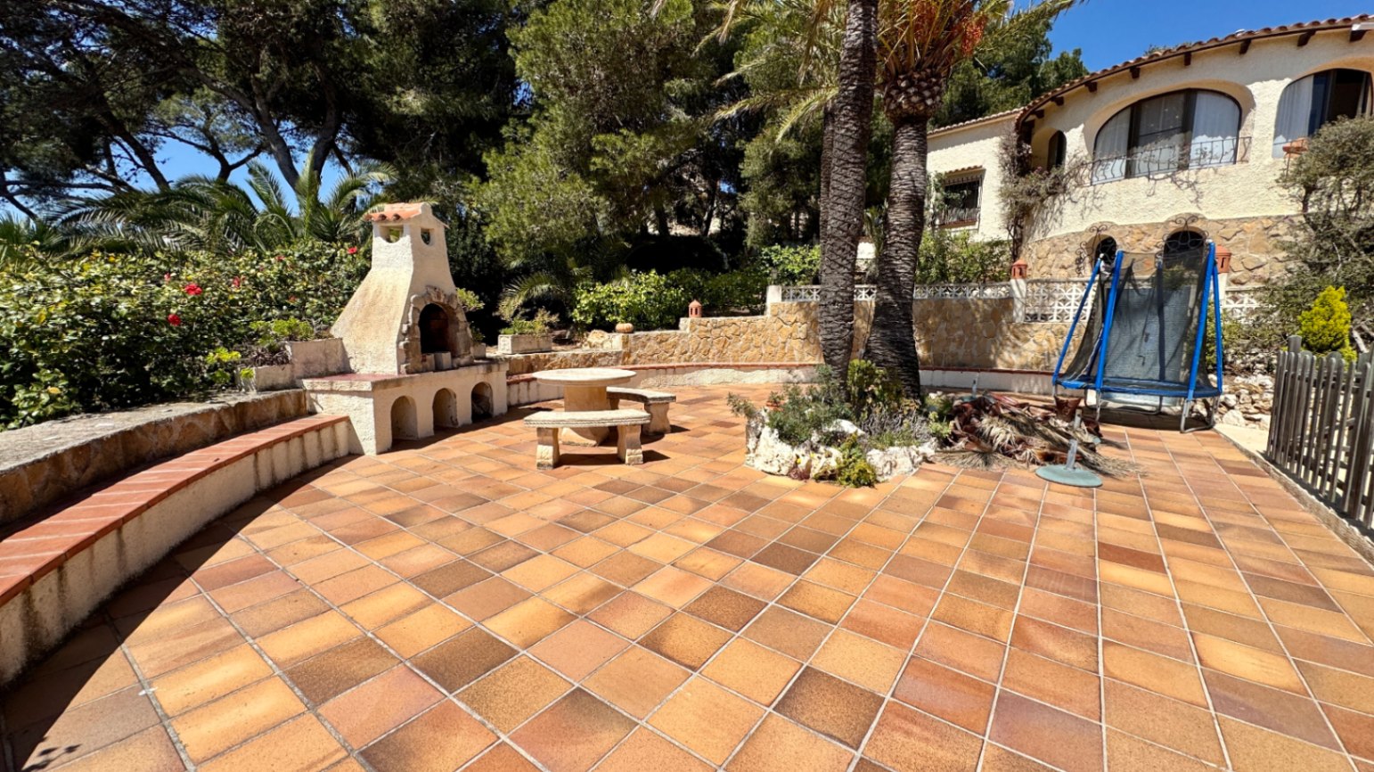 Ruim terras met terracotta tegels, een ingebouwde stenen barbecue, stenen zitmeubilair en palmbomen bij een mediterrane villa.